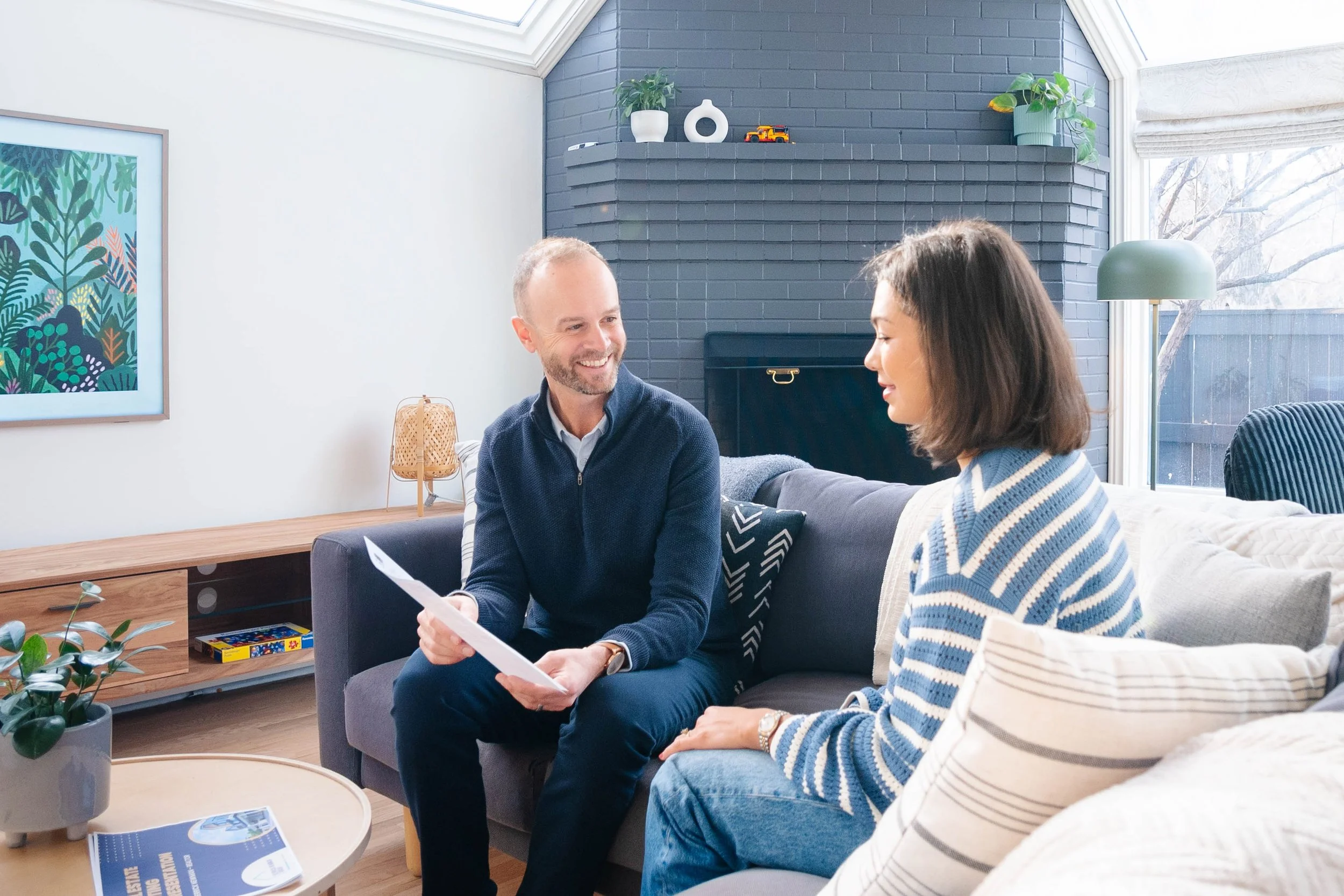 A man and a woman sitting on a sofa in a living room having a conversation, holding papers, with a window showing trees outside in the background.