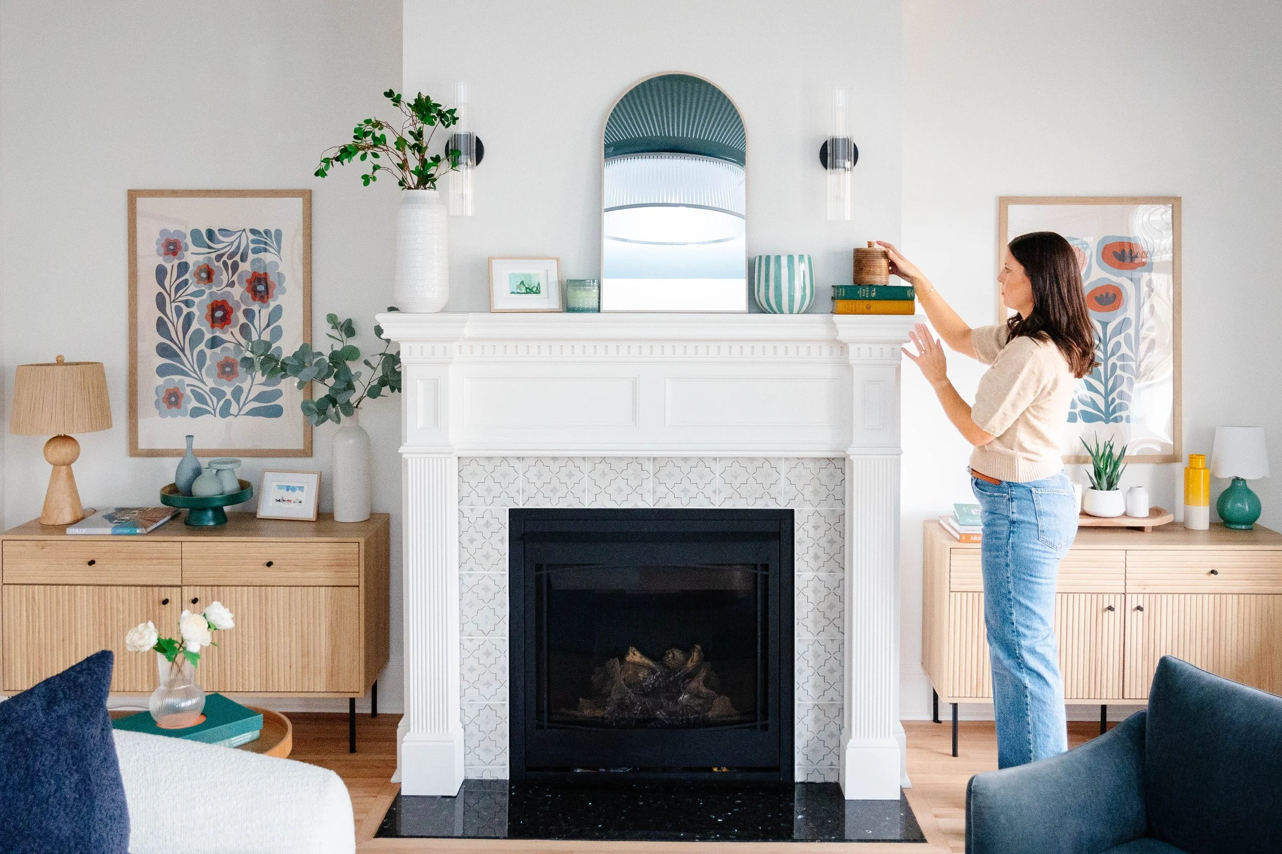 A woman places items on a shelf above a white fireplace mantel in a living room with light-colored walls, wooden furniture, and colorful decor.