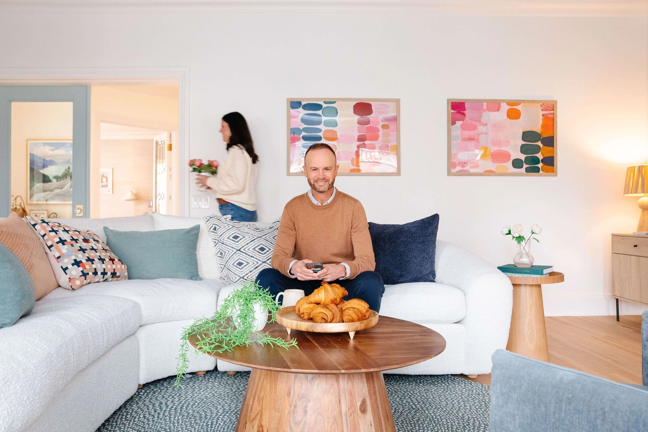 A man sitting on a white sectional sofa holding a remote control with a plate of croissants on the coffee table in front of him. A woman with flowers walks past in the background. The living room has colorful abstract art on the walls and a side table with a small vase of white flowers.