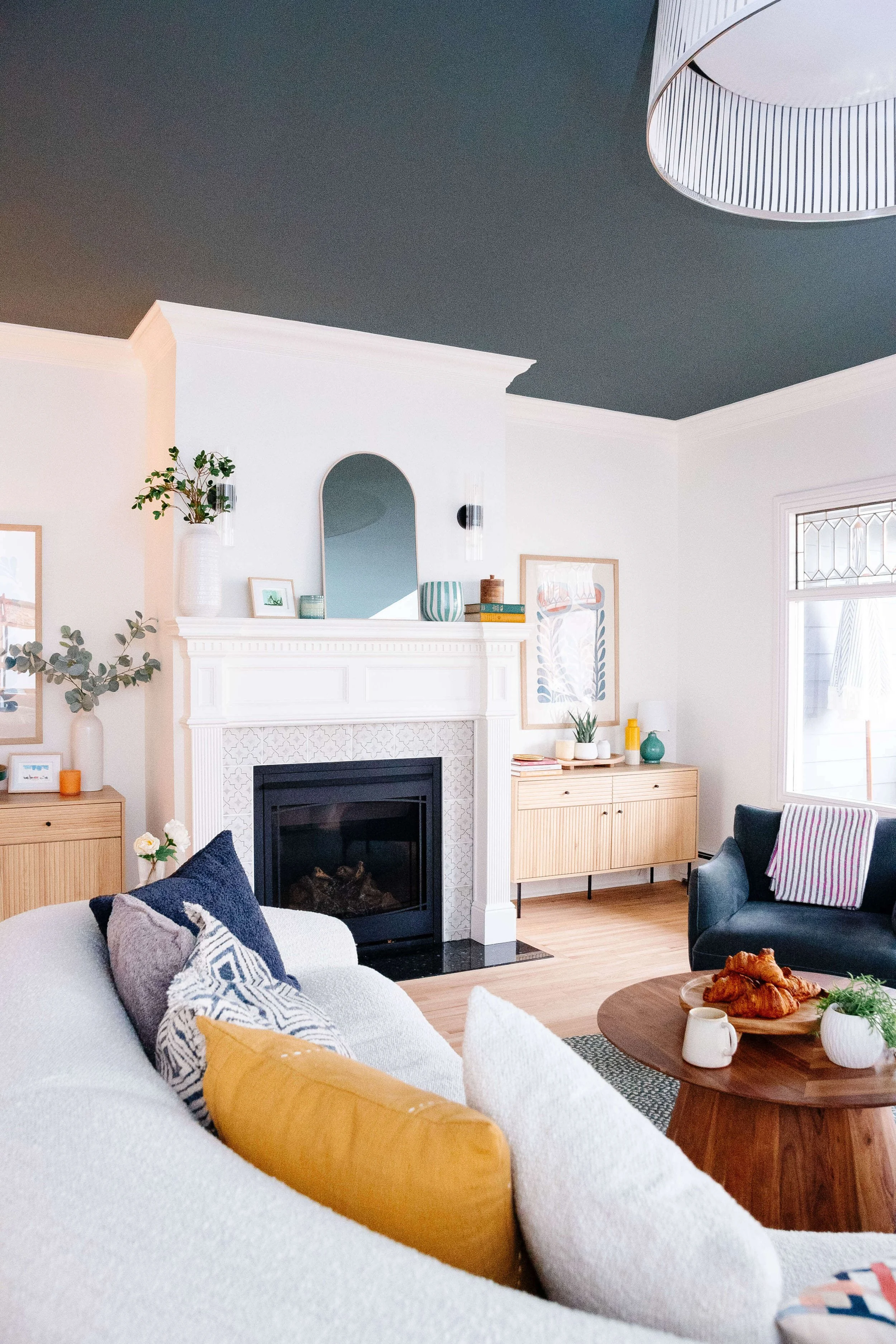 Living room with a black ceiling, white walls, white fireplace, and minimalistic decor. There is a white sofa with colorful pillows, a wooden coffee table with croissants and a mug, and a window allowing natural light.