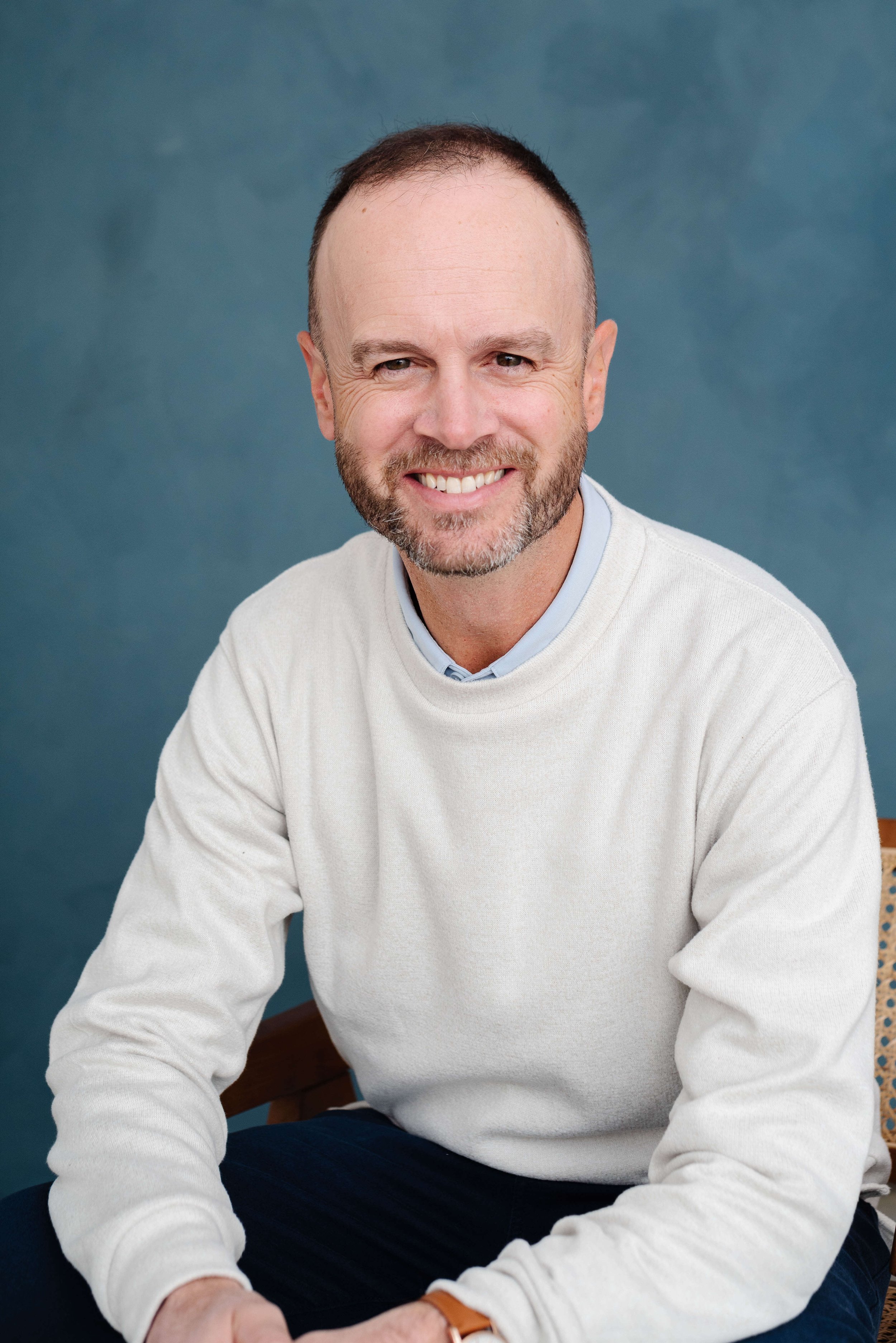 A smiling middle-aged man with short hair and a beard, wearing a white sweater and blue shirt, sitting in front of a blue background.