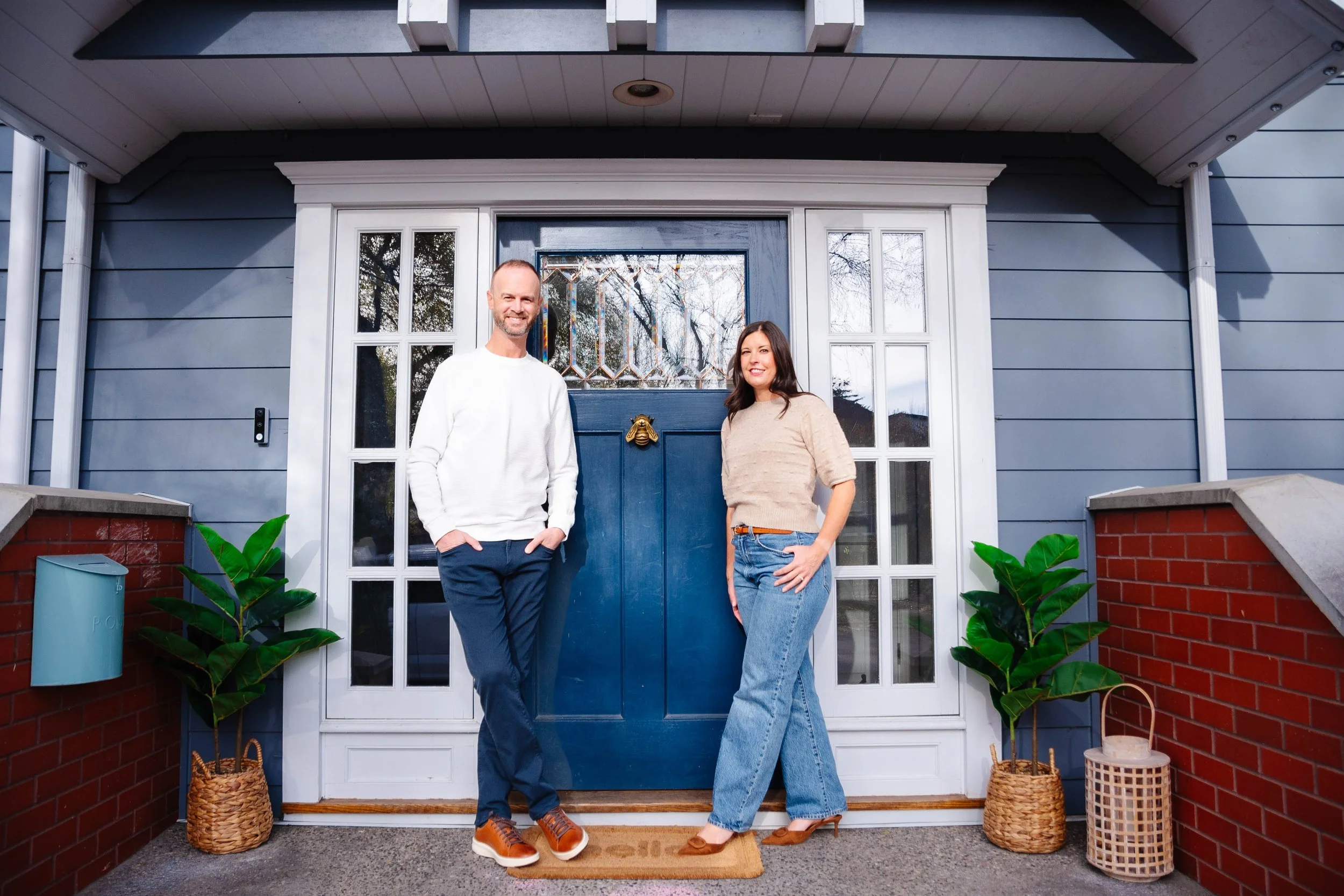 A man and a woman standing in front of a blue front door with glass panes, smiling. The man is wearing a white sweater, dark pants, and brown shoes. The woman is wearing a beige sweater, light blue jeans, and high heels. There are two potted plants on either side of the door, a mailbox on the left, and a small wicker basket on the right. The house has gray siding and red brick accents.
