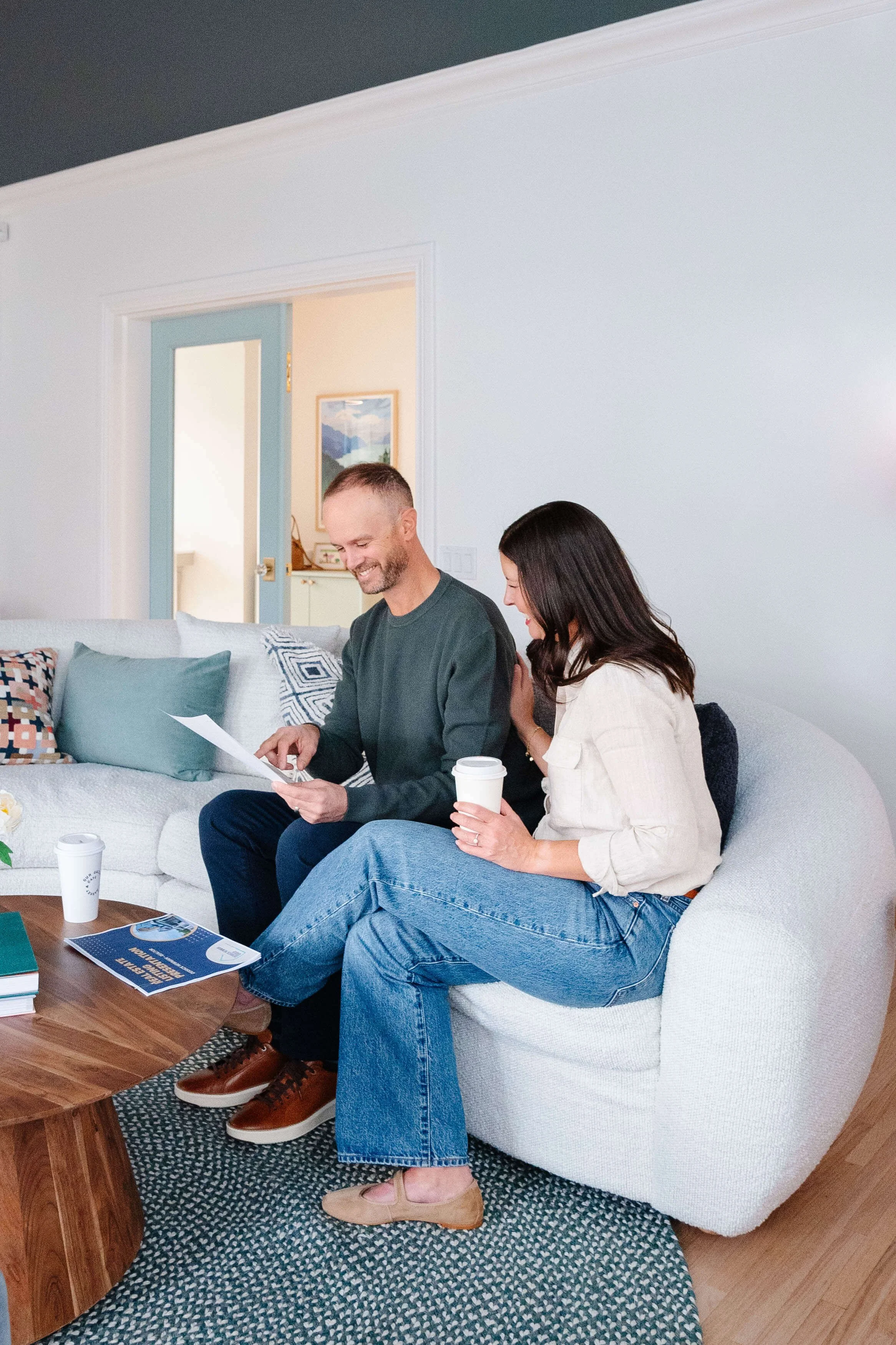 A man and woman sitting on a white couch, smiling, looking at papers together, each holding a coffee cup.