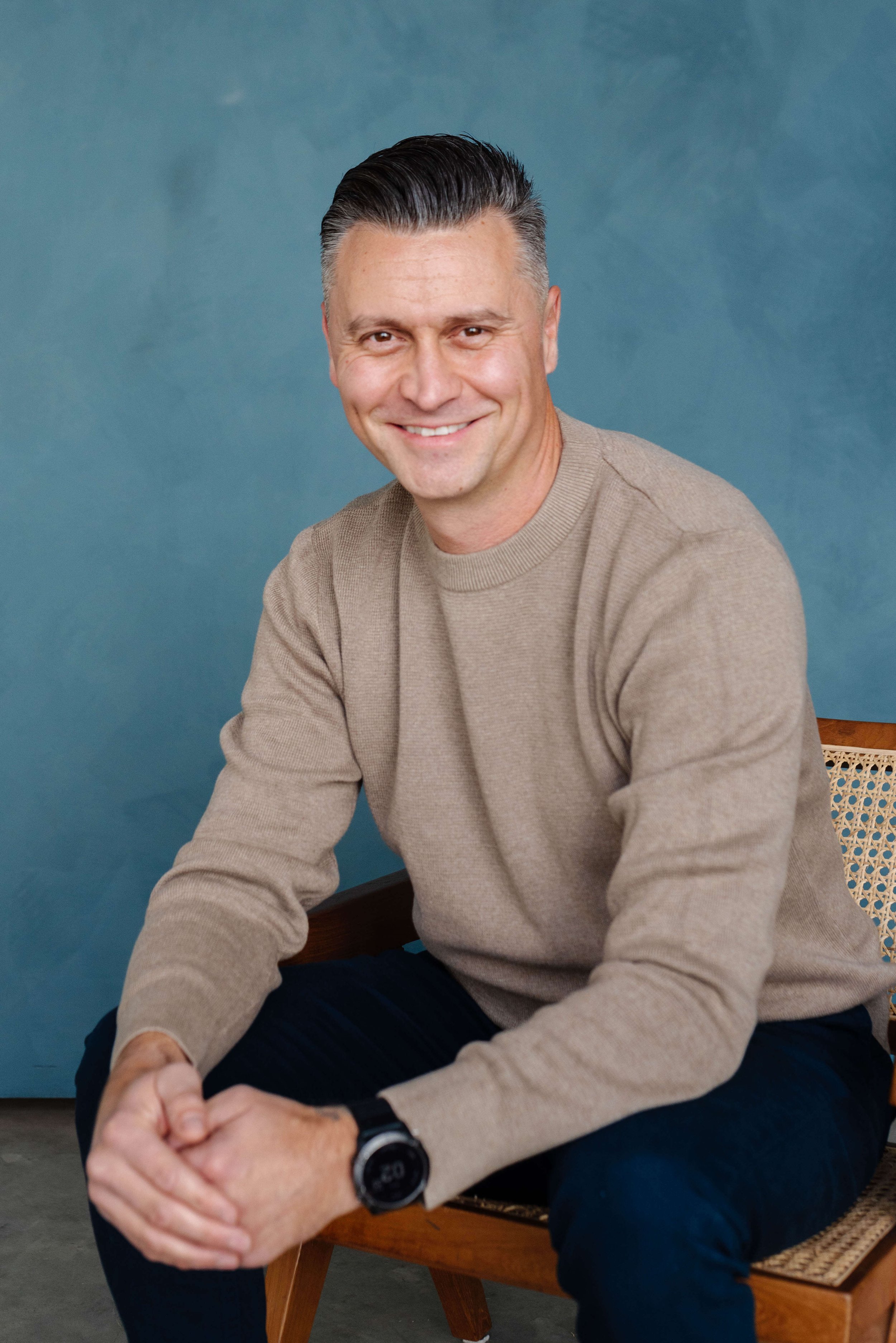 Smiling man with dark slicked-back hair, wearing a beige sweater, sitting on a wooden chair with a cane backrest against a blue textured background.