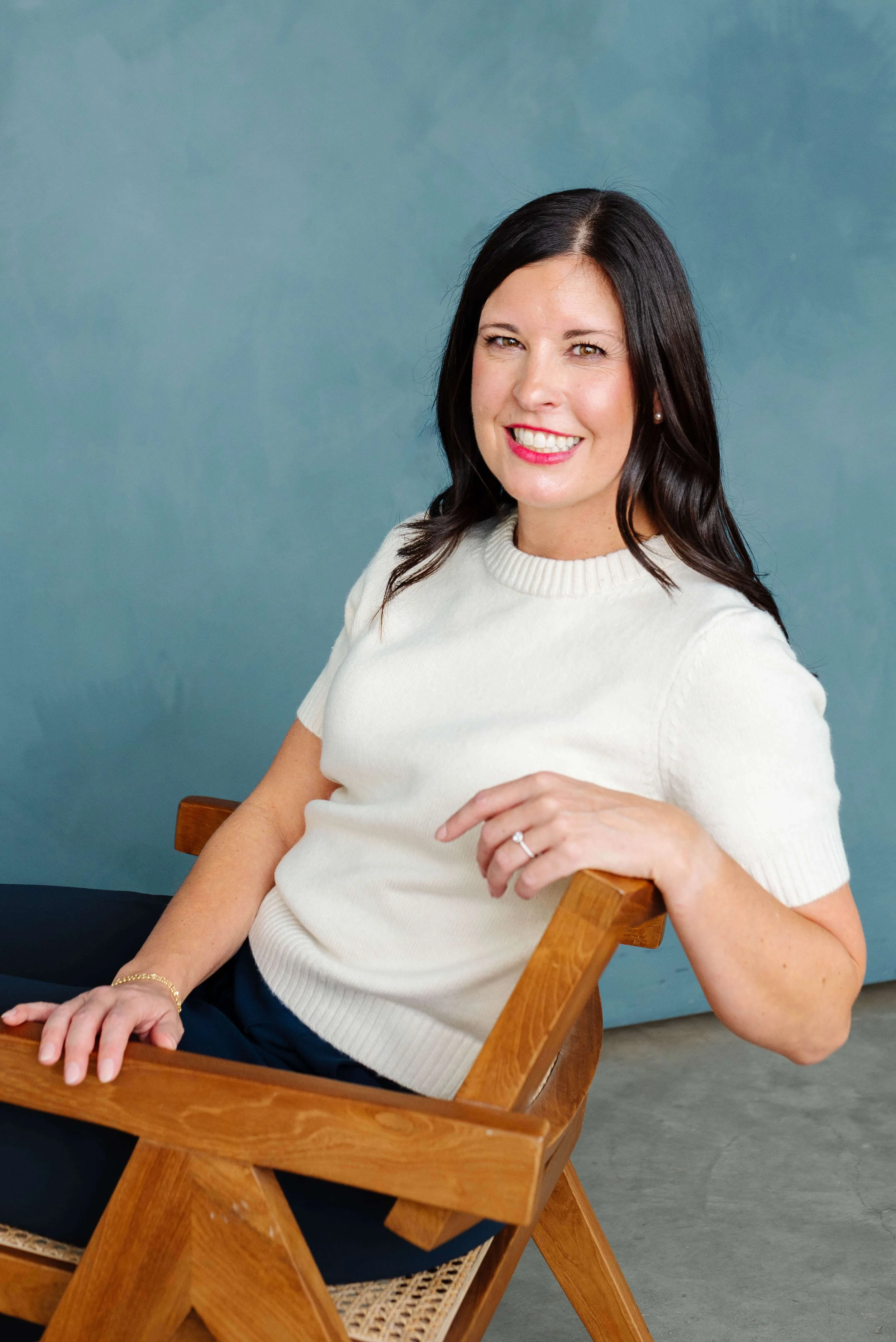 A woman with dark hair and a white sweater smiling at the camera, sitting on a wooden chair with a blue background.