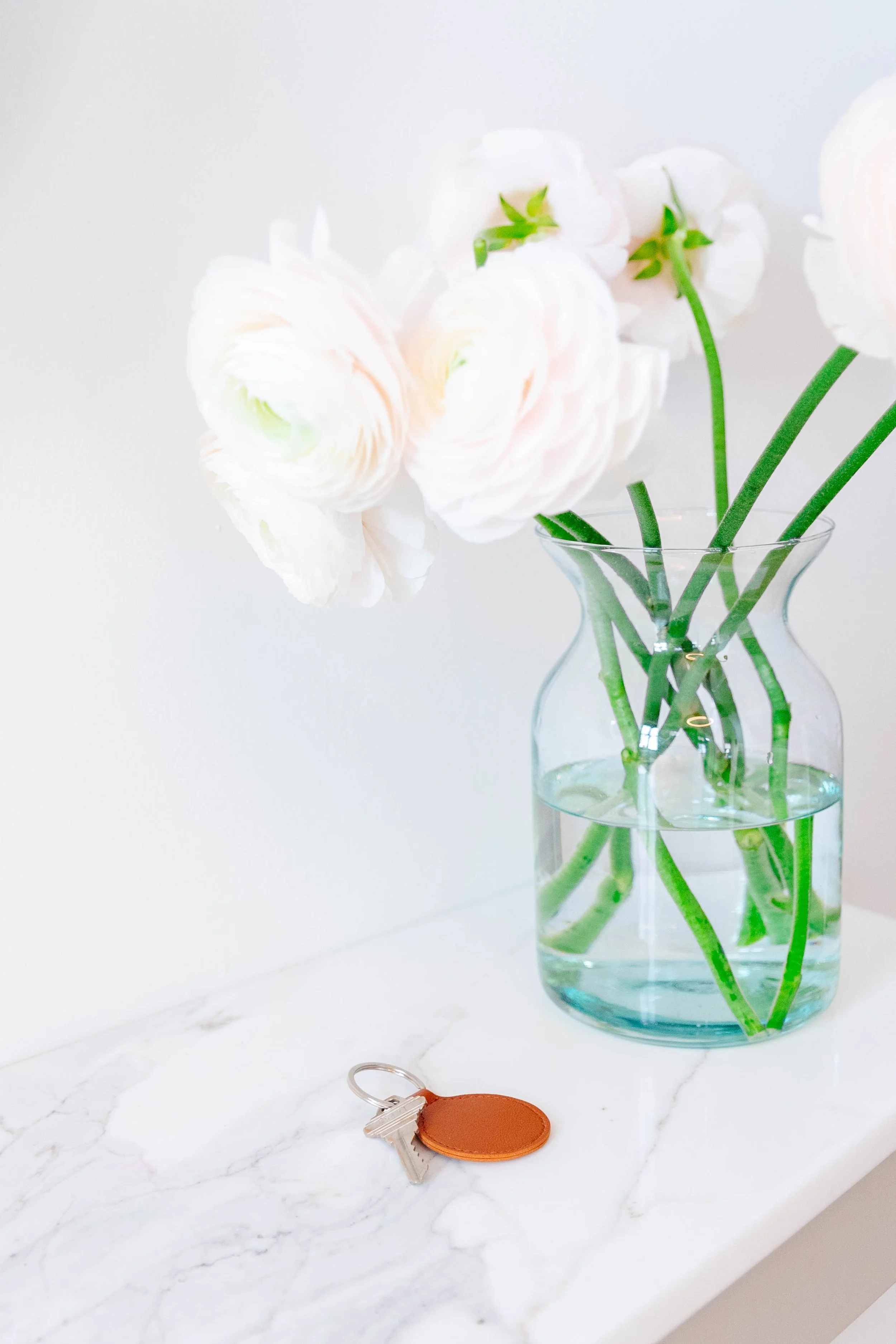 A glass vase with white and light pink ranunculus flowers and green stems, placed on a white marble surface. A small set of keys with a brown leather keychain is on the marble surface near the vase.