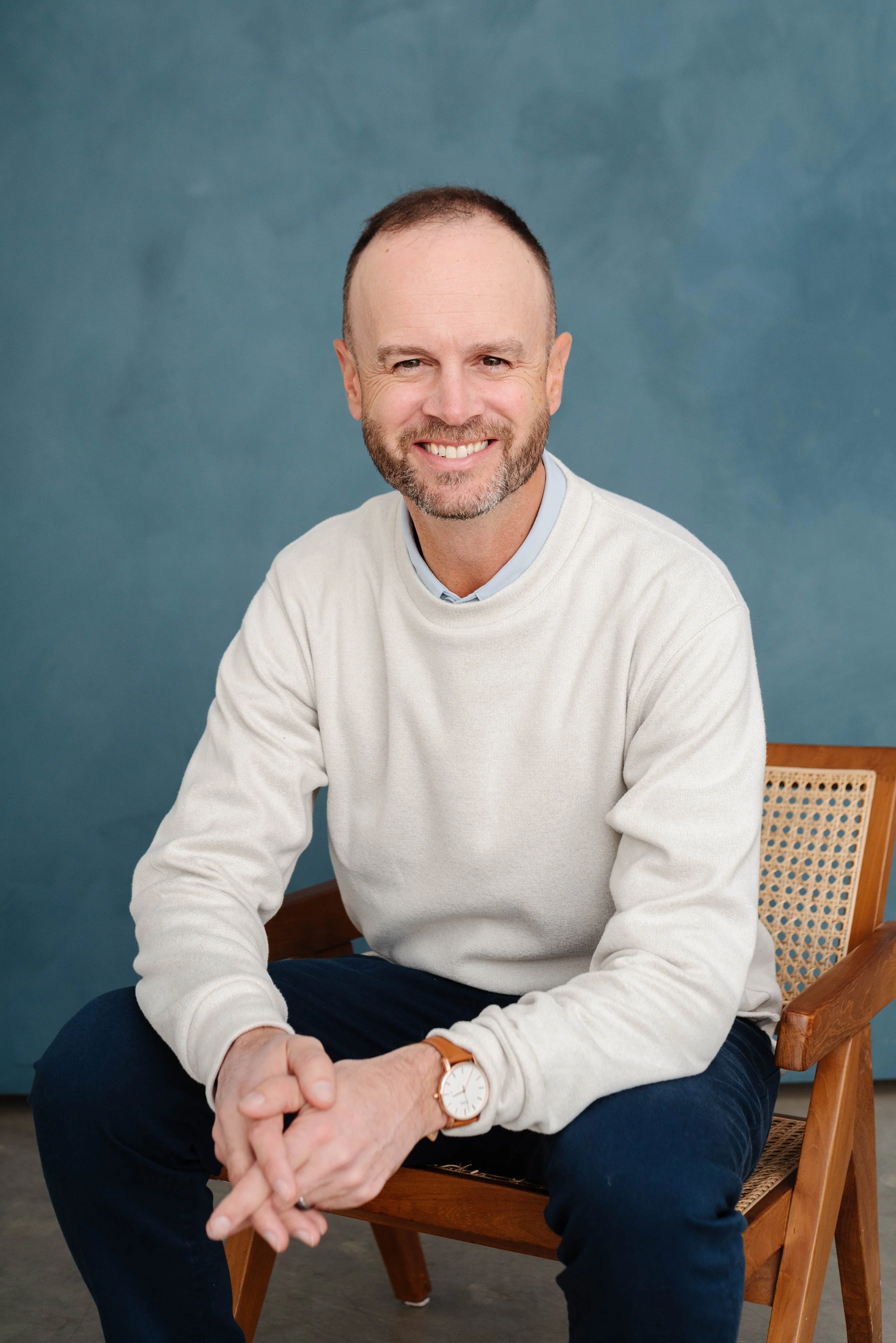 A smiling man with a beard sitting on a wooden chair, wearing a white sweater, blue jeans, and a watch, against a blue-green background.