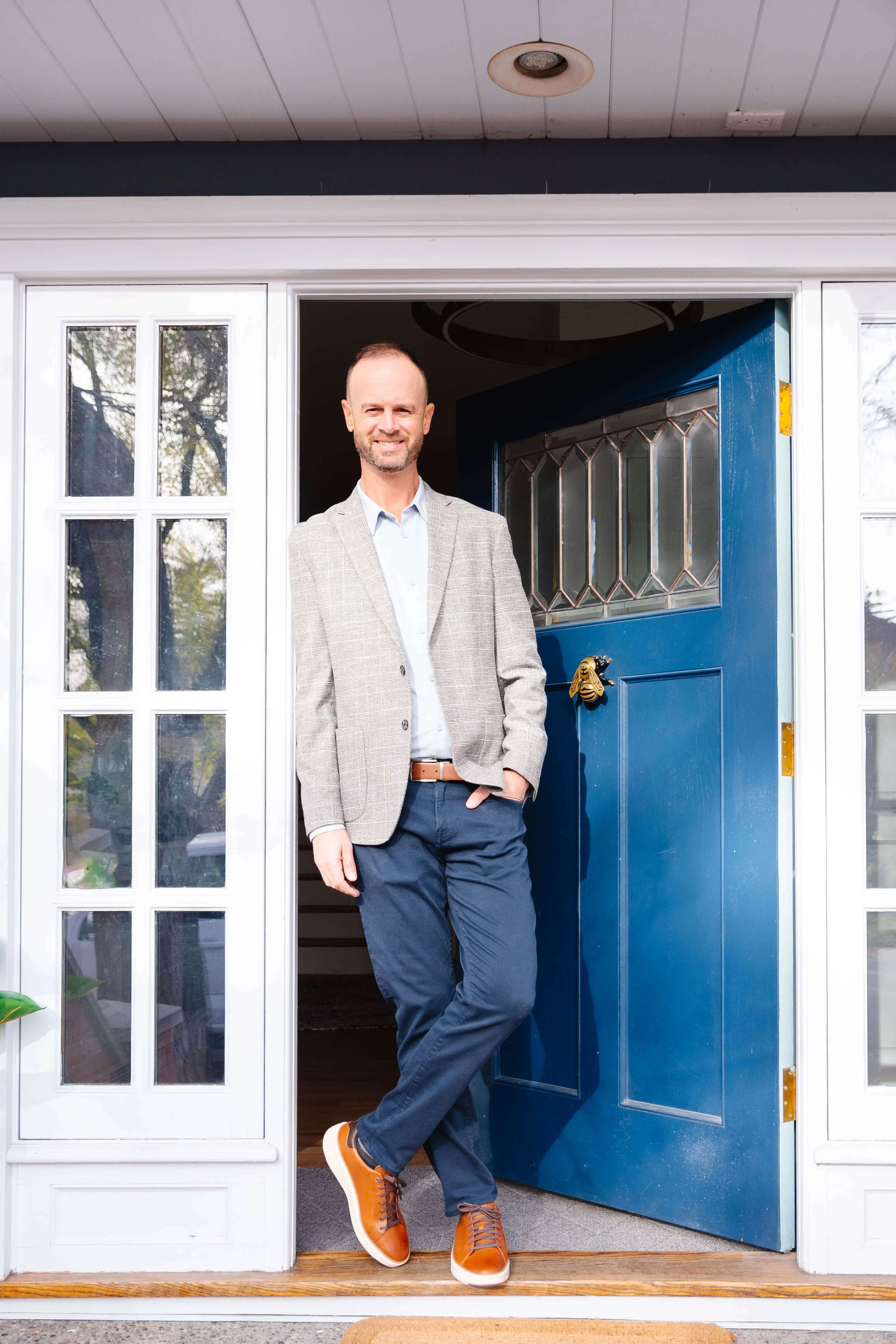 A man standing at the open front door of a house, smiling, wearing a light gray blazer, light blue shirt, navy blue pants, and brown sneakers.