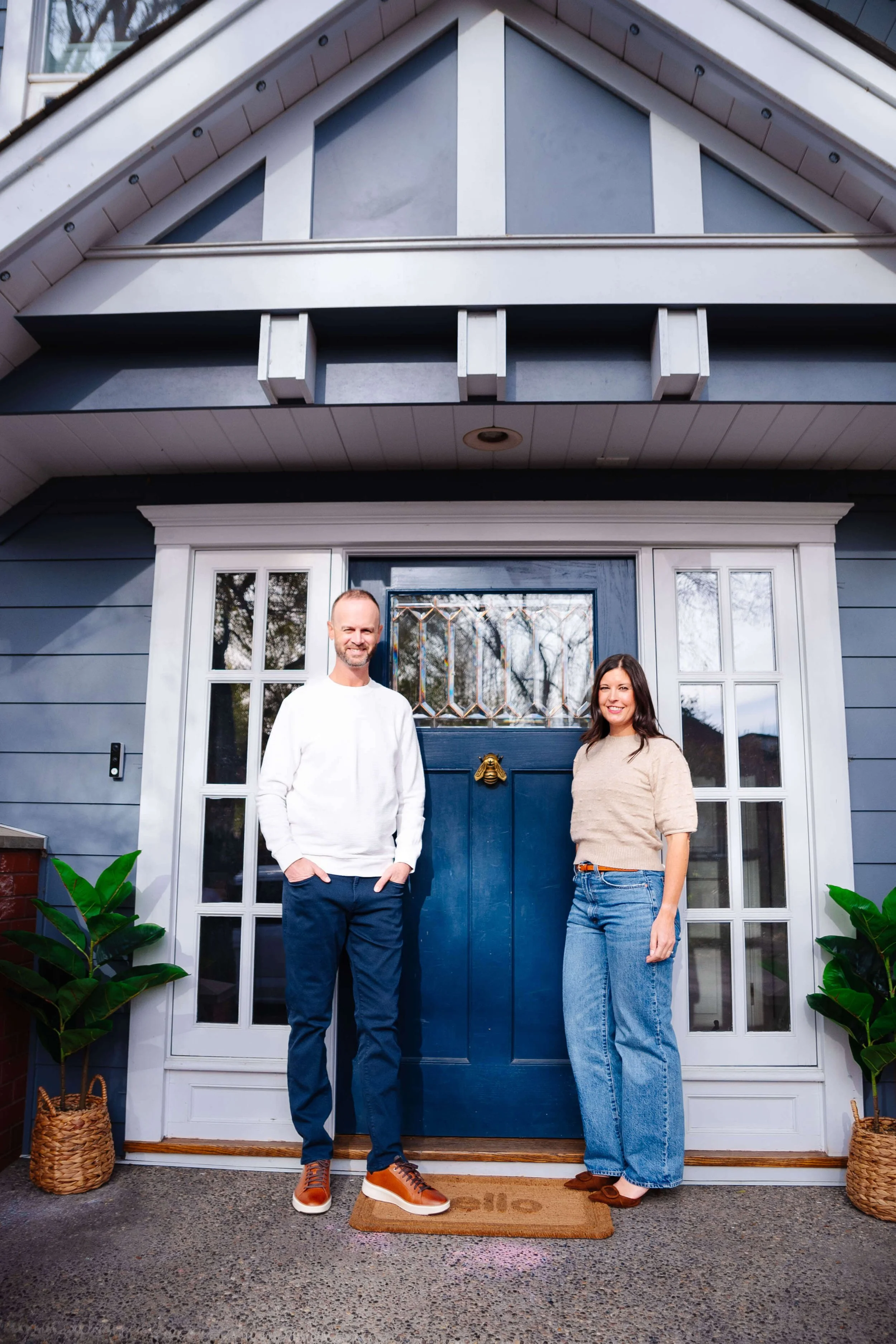 A man and woman standing in front of a house door with white trim and blue walls, smiling. The man is wearing a white sweater and blue pants, and the woman is wearing a beige sweater and blue jeans. There are potted plants on either side of the door and a Welcome mat on the ground.
