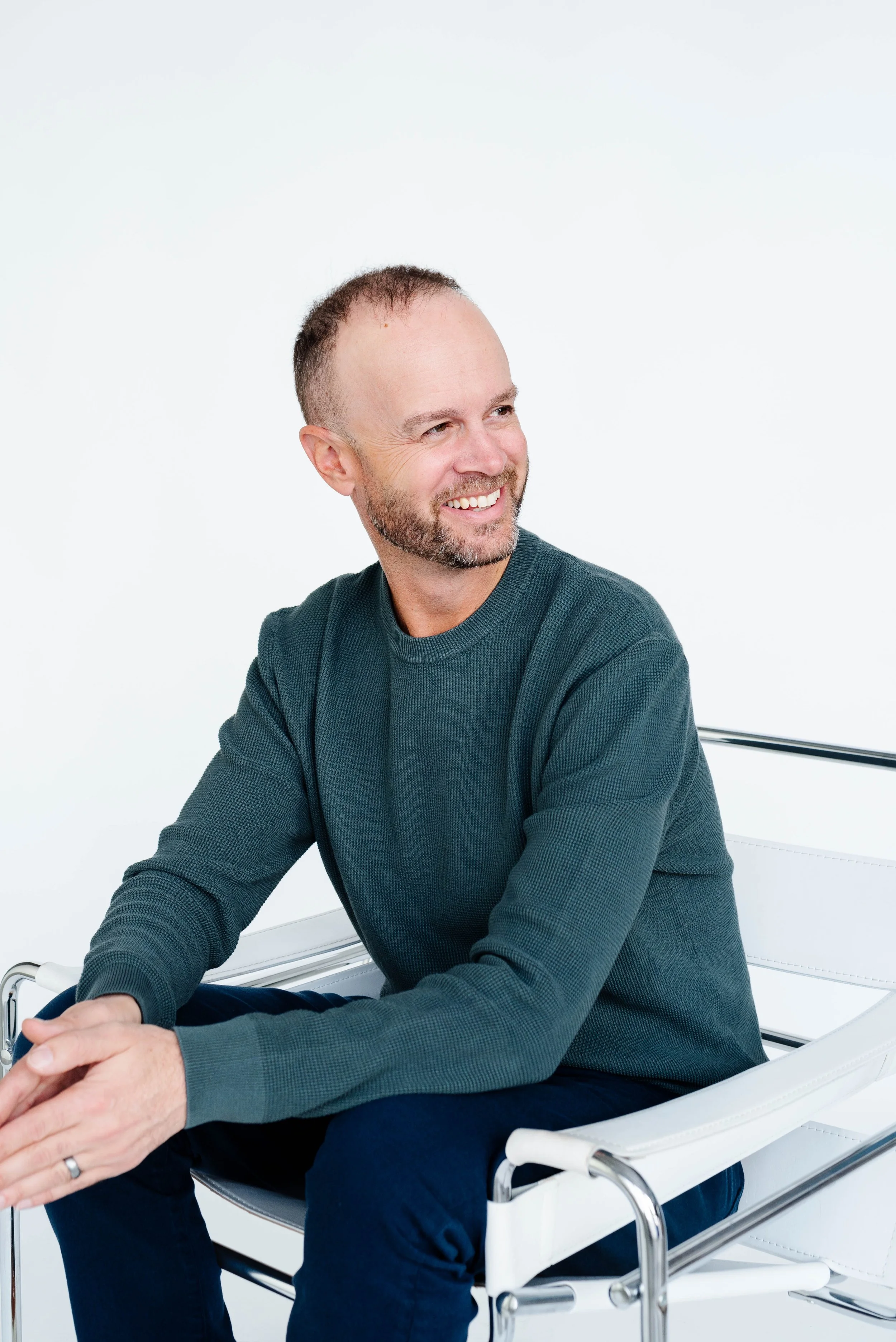 A smiling middle-aged man with a short beard and receding hair, sitting on a modern white chair against a plain white background.