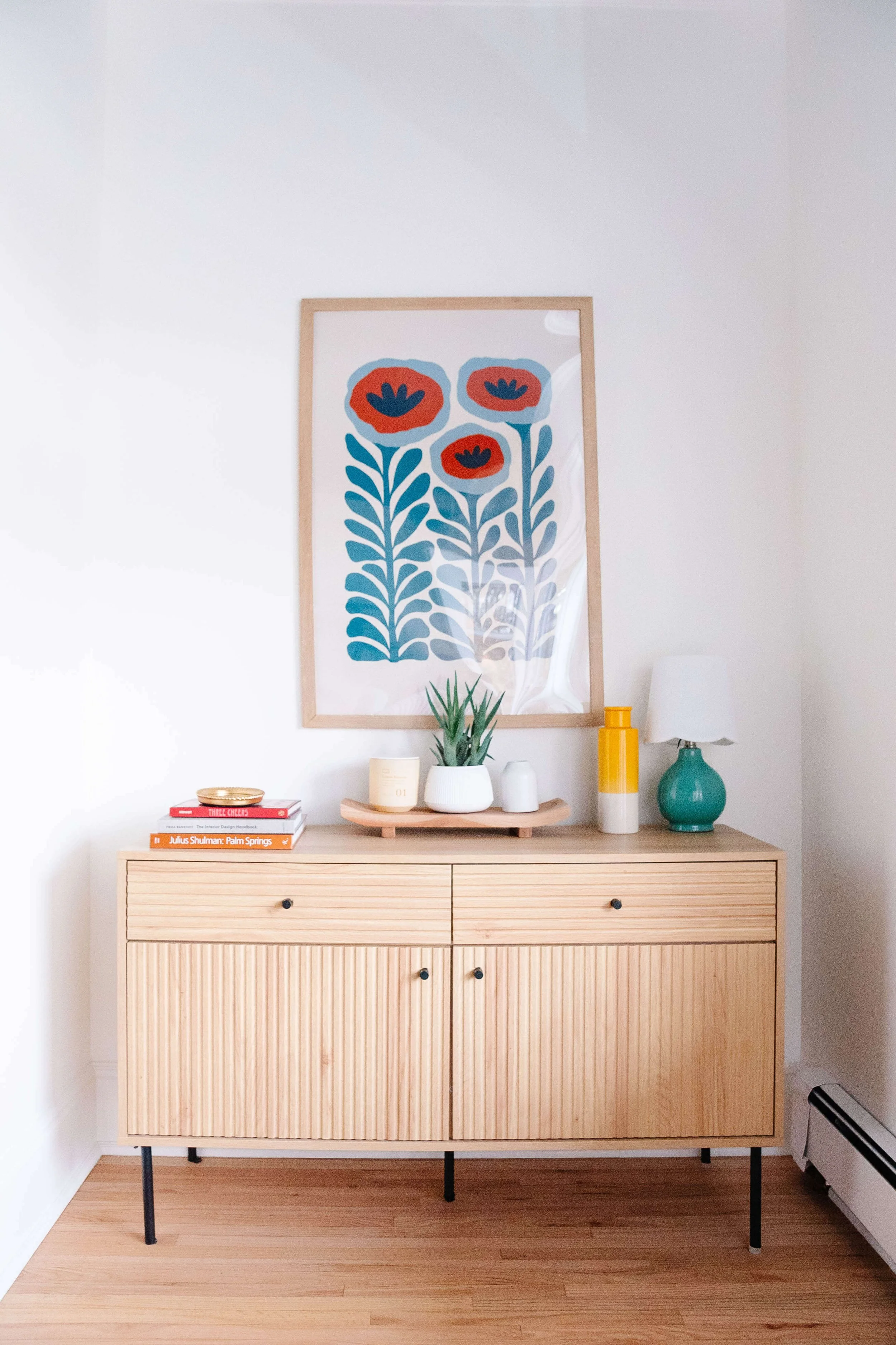 A wooden sideboard with a potted plant, books, a small vase, a yellow and gray vase, and a teal lamp on top, with a floral artwork hanging above it.