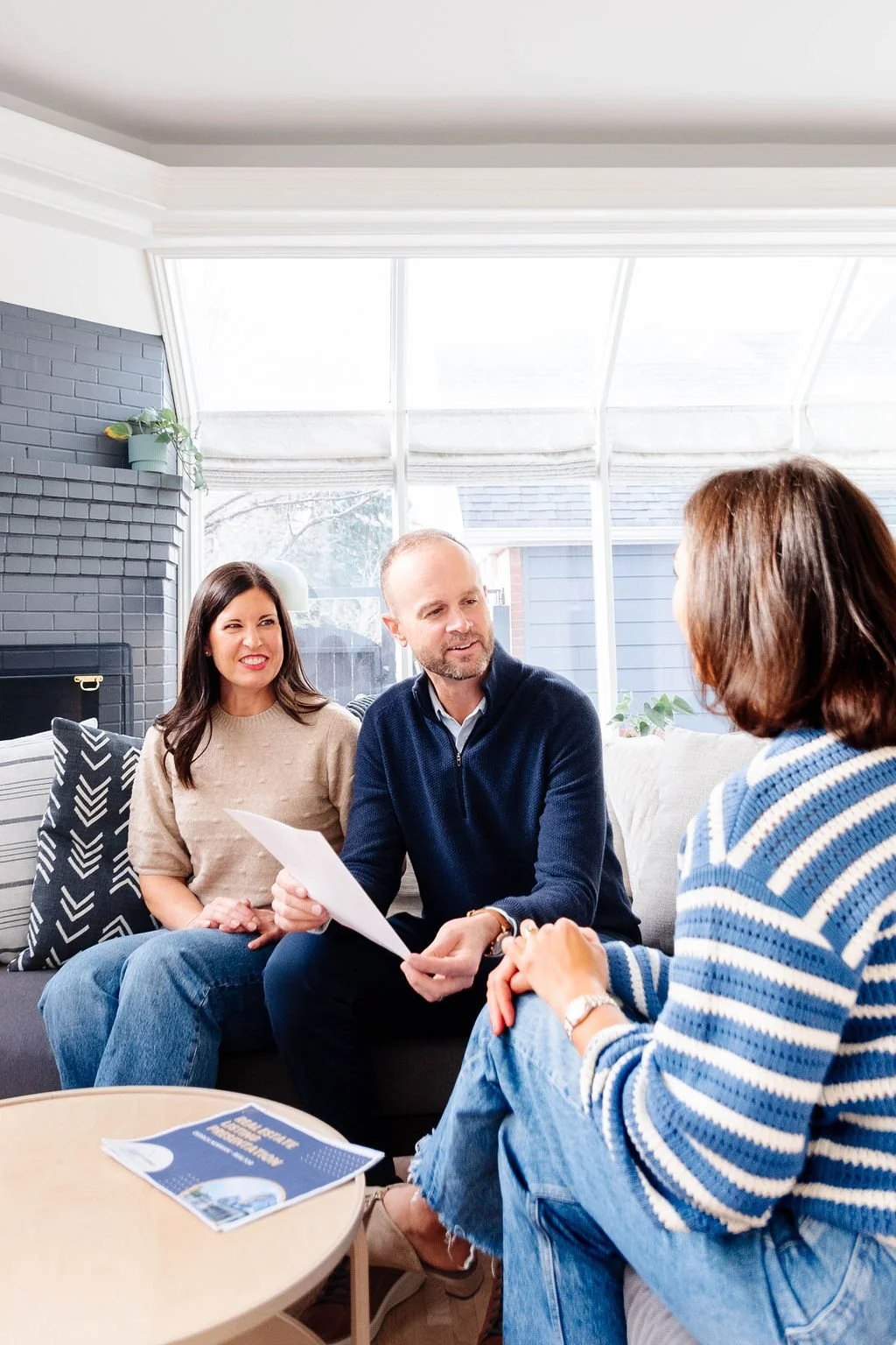Three adults sitting on a sofa in a bright living room engaged in a discussion, with a woman facing away wearing a blue striped sweater, a man in the middle holding a paper, and a woman on the left smiling.