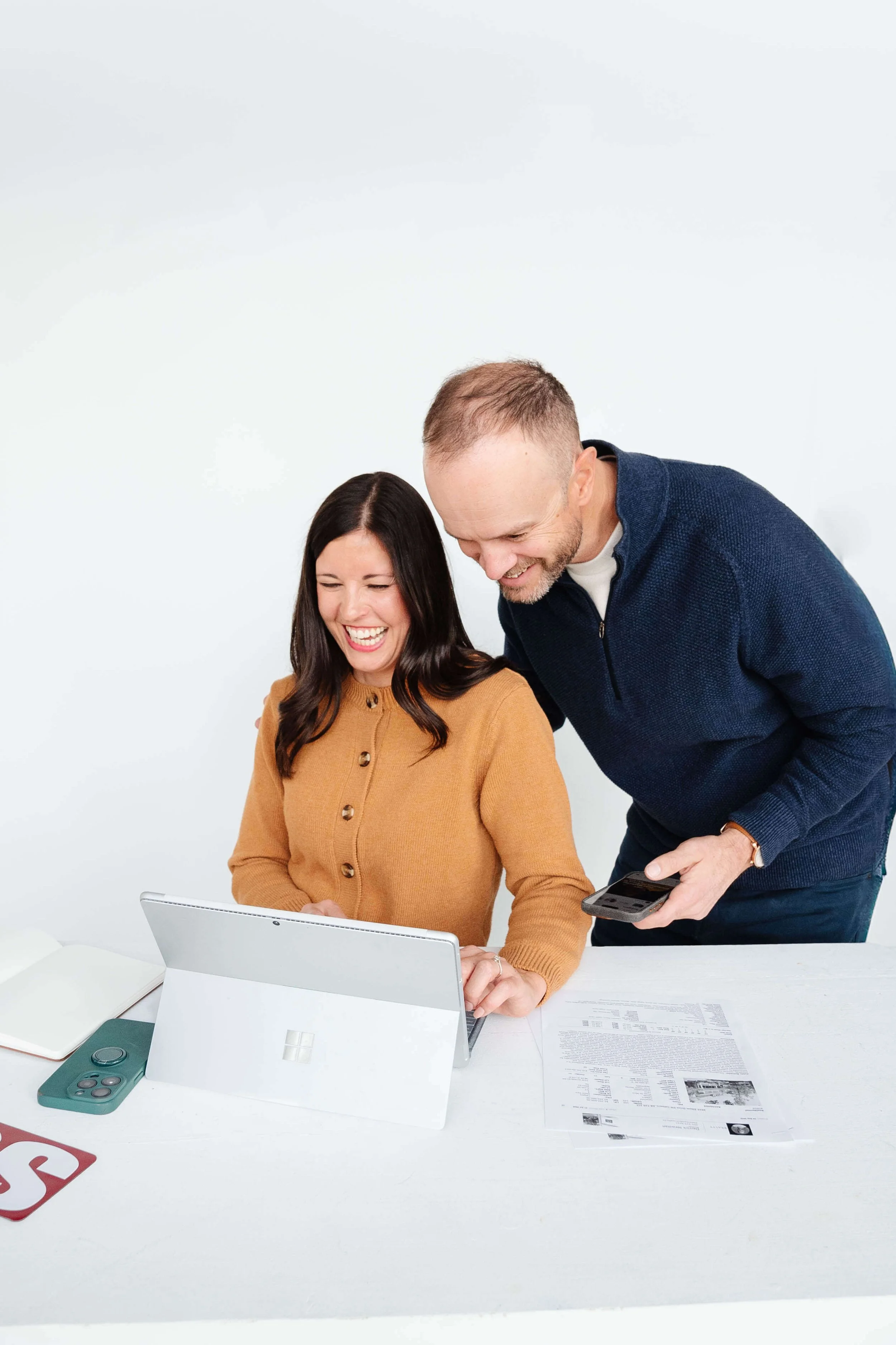 A man and woman sitting and standing at a white table, laughing and looking at a laptop and smartphone, with papers and remote controls on the table.