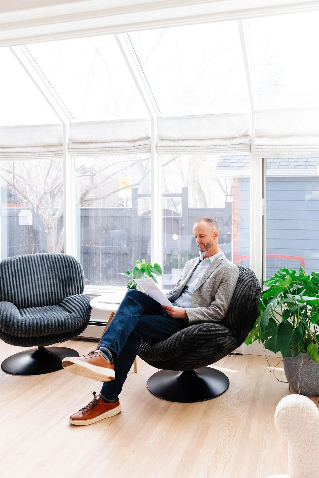 A man in a gray blazer and blue jeans sitting on a black, round chair reading a document in a bright sunlit room with large windows and green plants.
