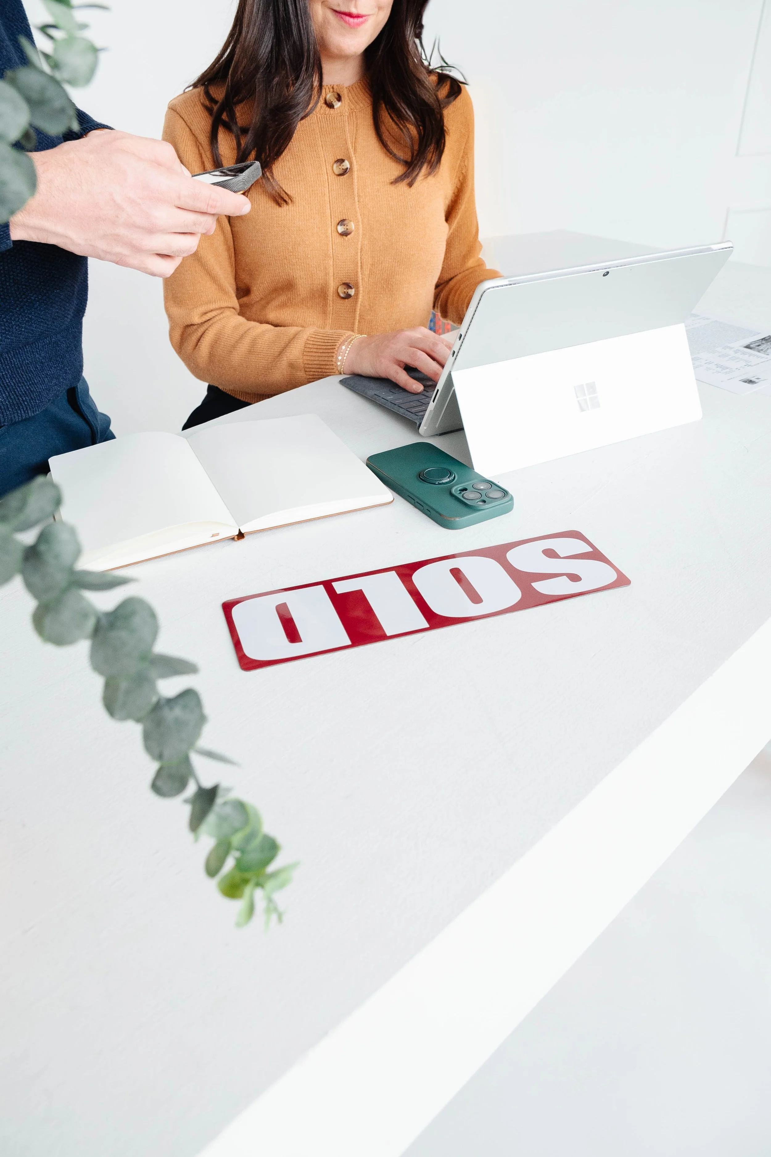 A woman working on a Surface tablet at a white desk with an open notebook, a smartphone, and a 'STOP' sign made of red and white blocks. A person's hand holding a phone appears partially in the frame, and a plant branch is visible in the foreground.