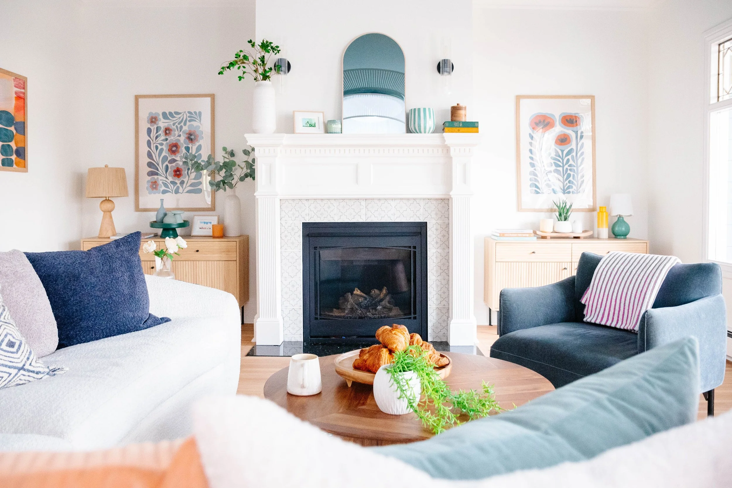 Bright living room with white walls, a white fireplace with decorative tile, framed floral artwork, a large mirror, potted plants, side tables with lamps, and comfortable seating including a white couch with navy and patterned pillows and a blue armchair with a striped cushion. A wooden coffee table holds croissants, a white vase with greenery, and a small cup.