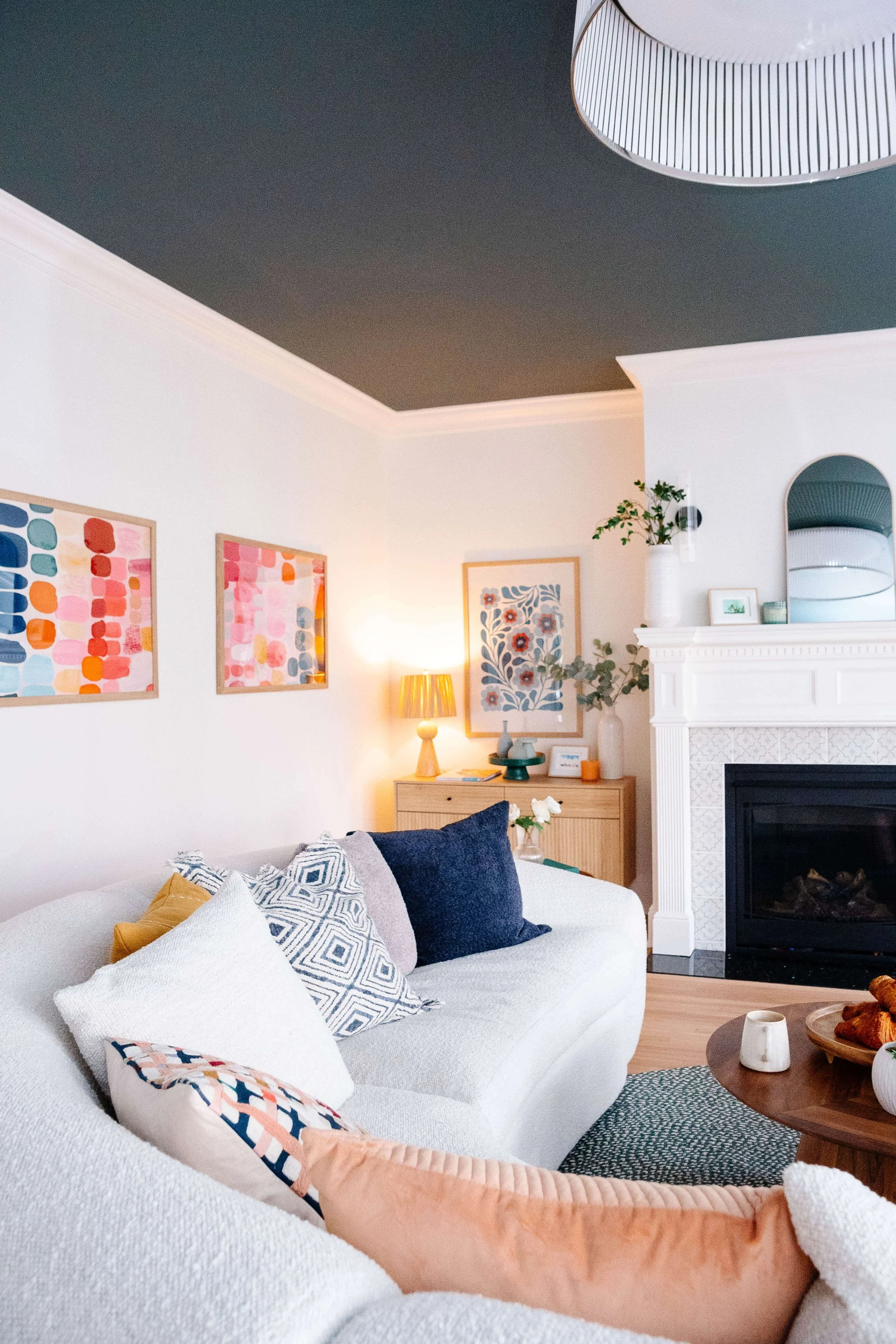 Living room with white sofa, colorful throw pillows, artwork on white walls, wooden side table with lamp and decorative items, a white fireplace with greenery and framed picture, and a dark ceiling with a hanging lamp.