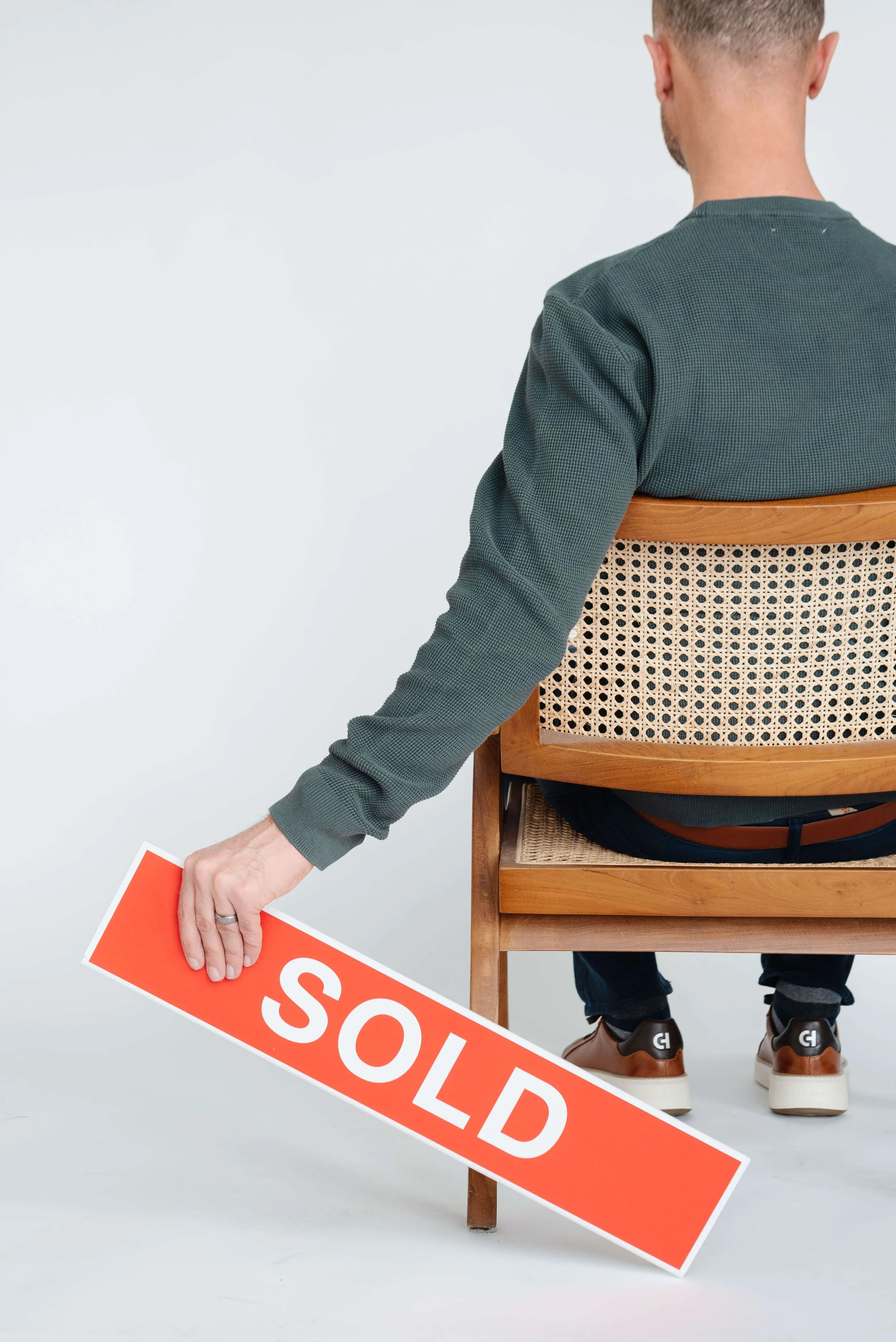 Man sitting on a wooden chair holding a red 'SOLD' sign in his left hand, facing away from the camera, against a plain white background.