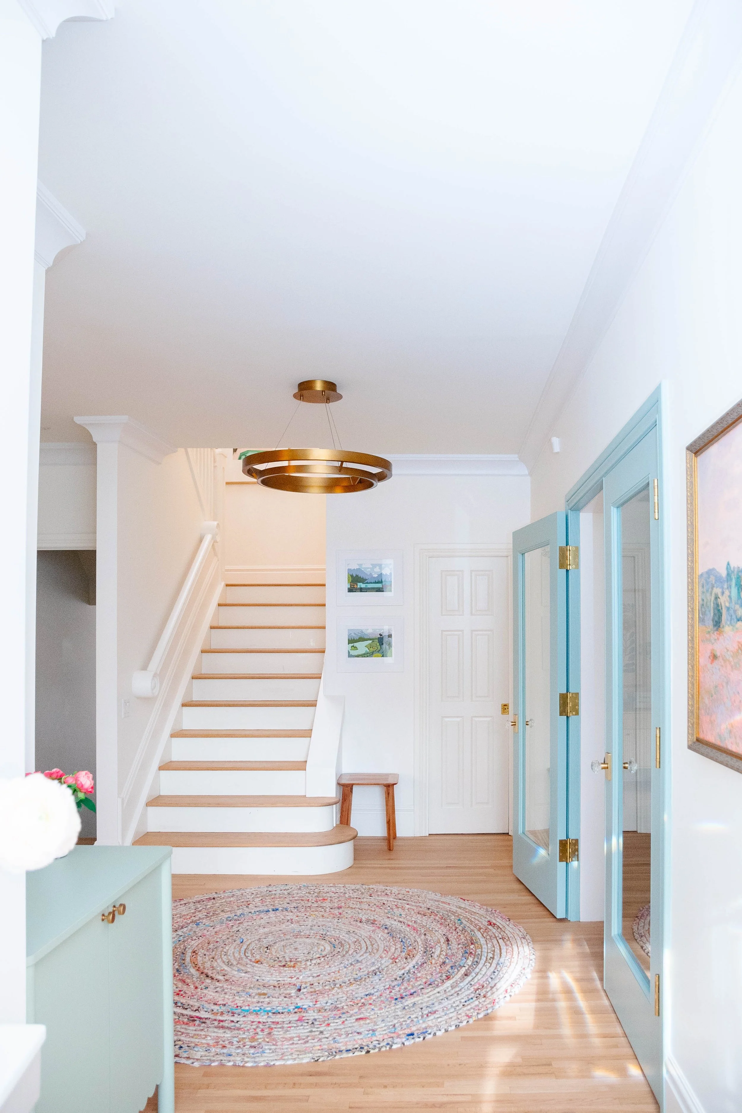 Bright entryway with a circular rug, staircase, blue doors with brass hinges, and artwork on white walls.