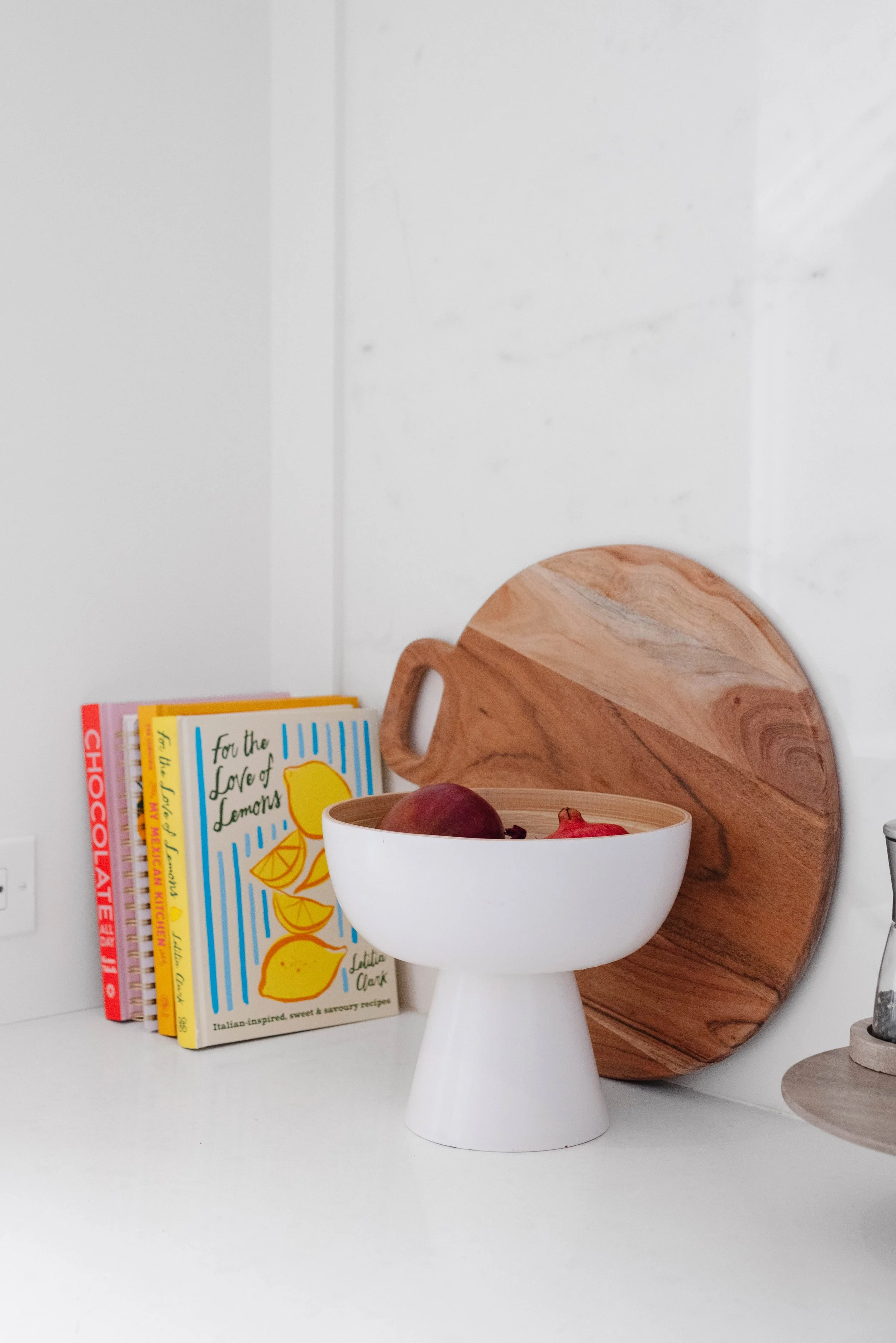 A white pedestal bowl filled with figs and pomegranates on a white kitchen countertop. Behind the bowl, a round wooden cutting board and a stack of colorful cookbooks.