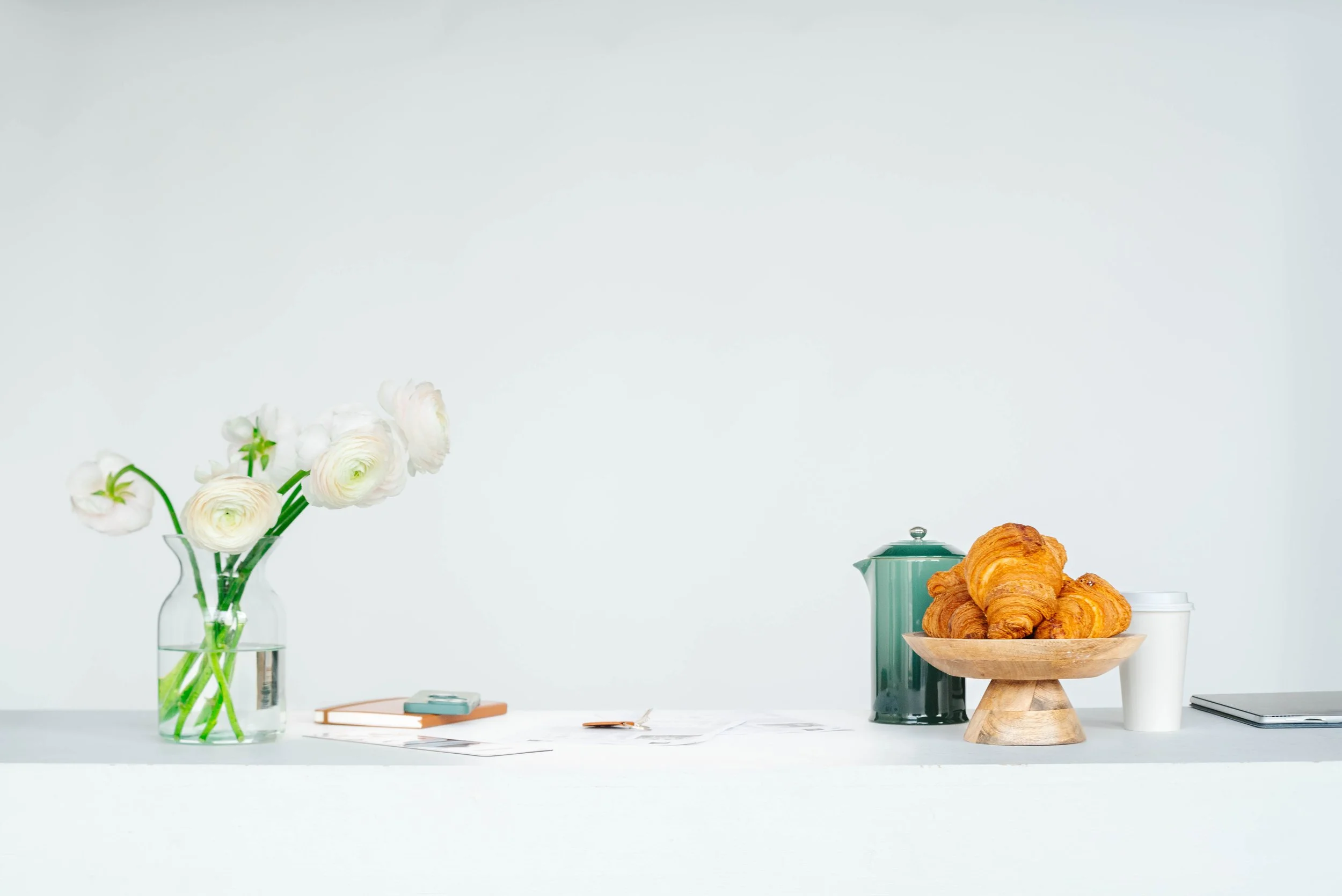 A minimalist table with a vase of white flowers, a notebook, a spoon, a green kettle, a wooden plate of croissants, a white cup, and a closed laptop against a plain white background.