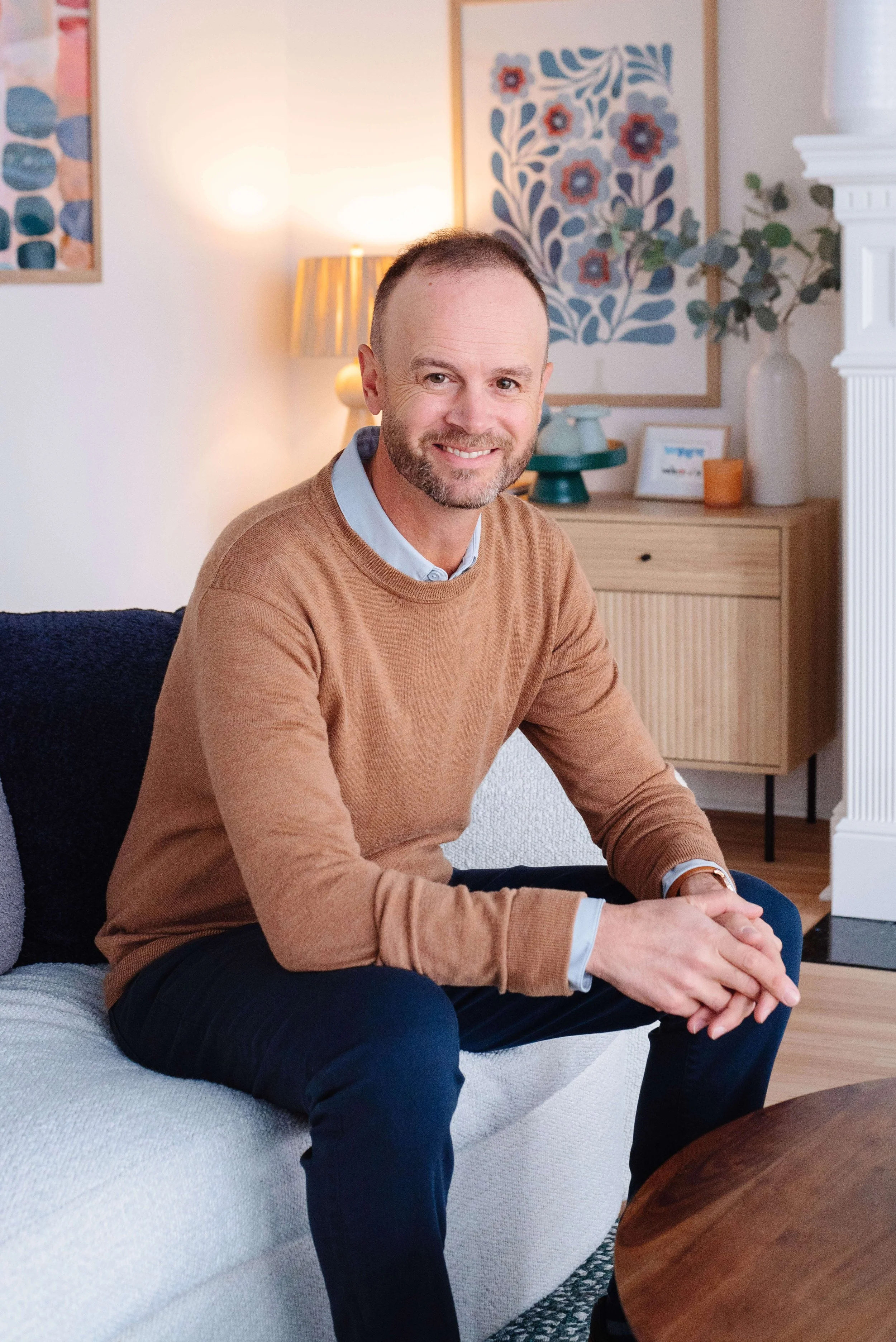 A middle-aged man with a beard and short hair, wearing a brown sweater and dark pants, sitting on a white couch in a living room with artwork and a lamp in the background.