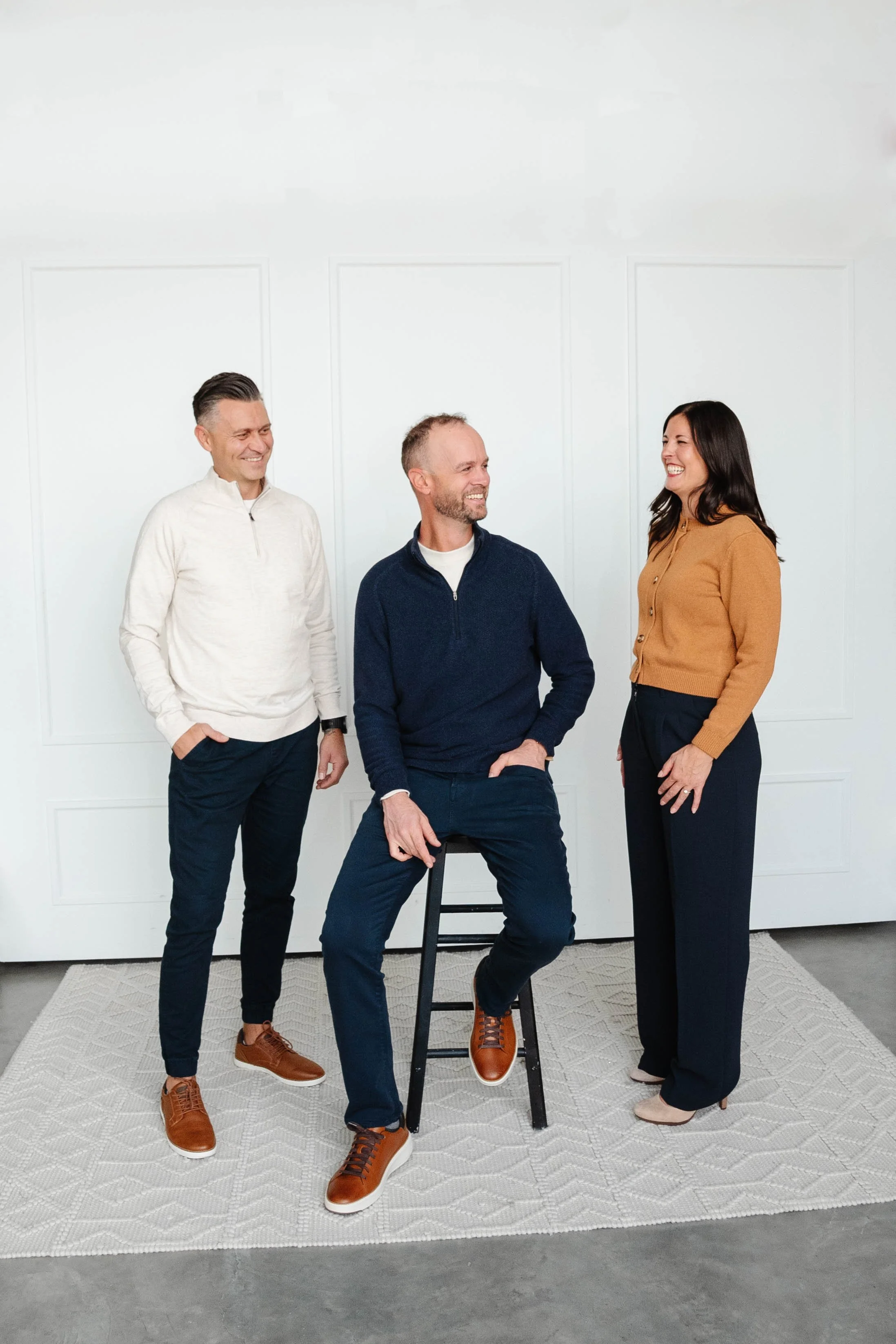 Three people smiling and talking in a room with white walls, one sitting on a black stool and the other two standing.