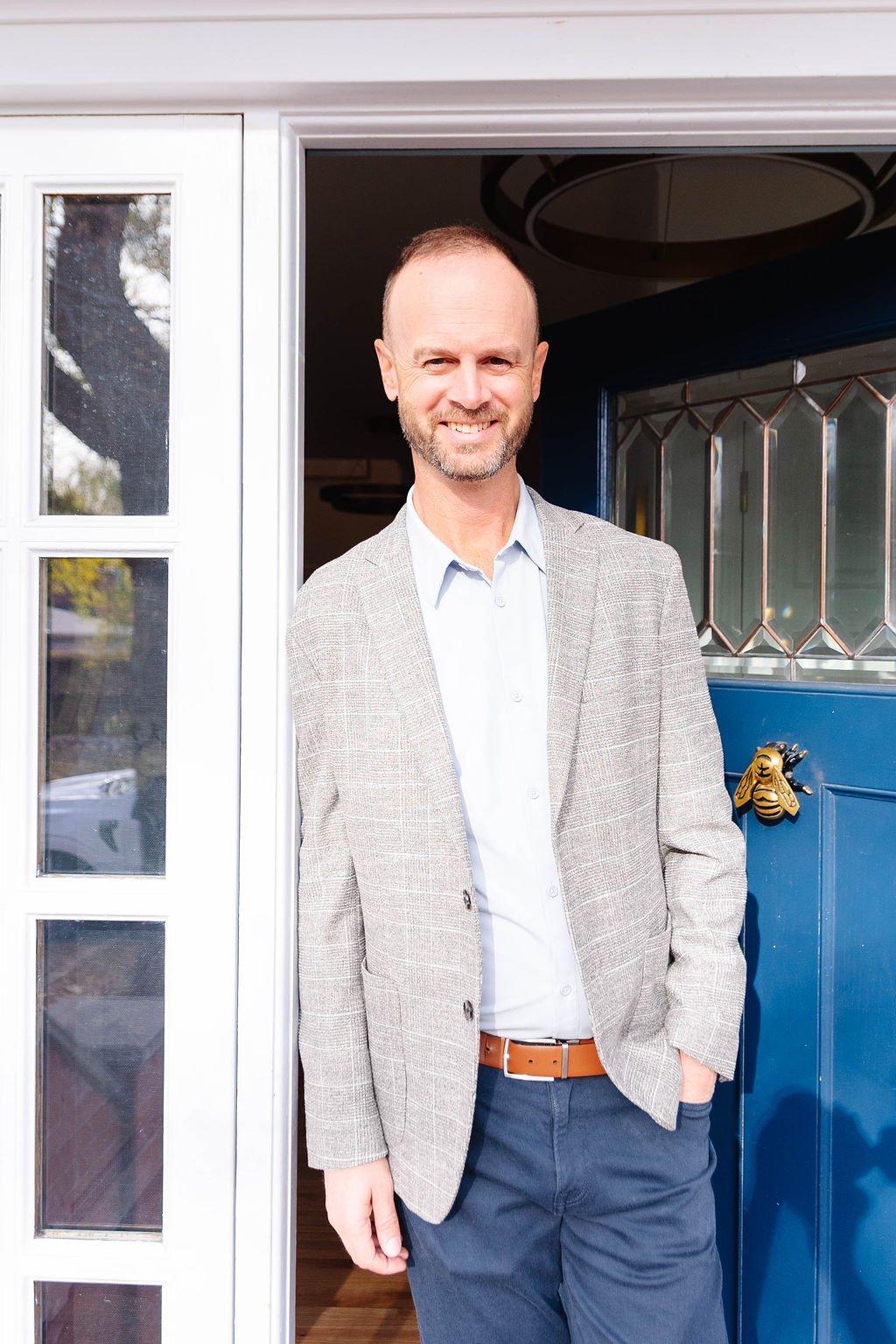 A smiling man with a beard wearing a gray blazer and light blue shirt standing at his doorway.