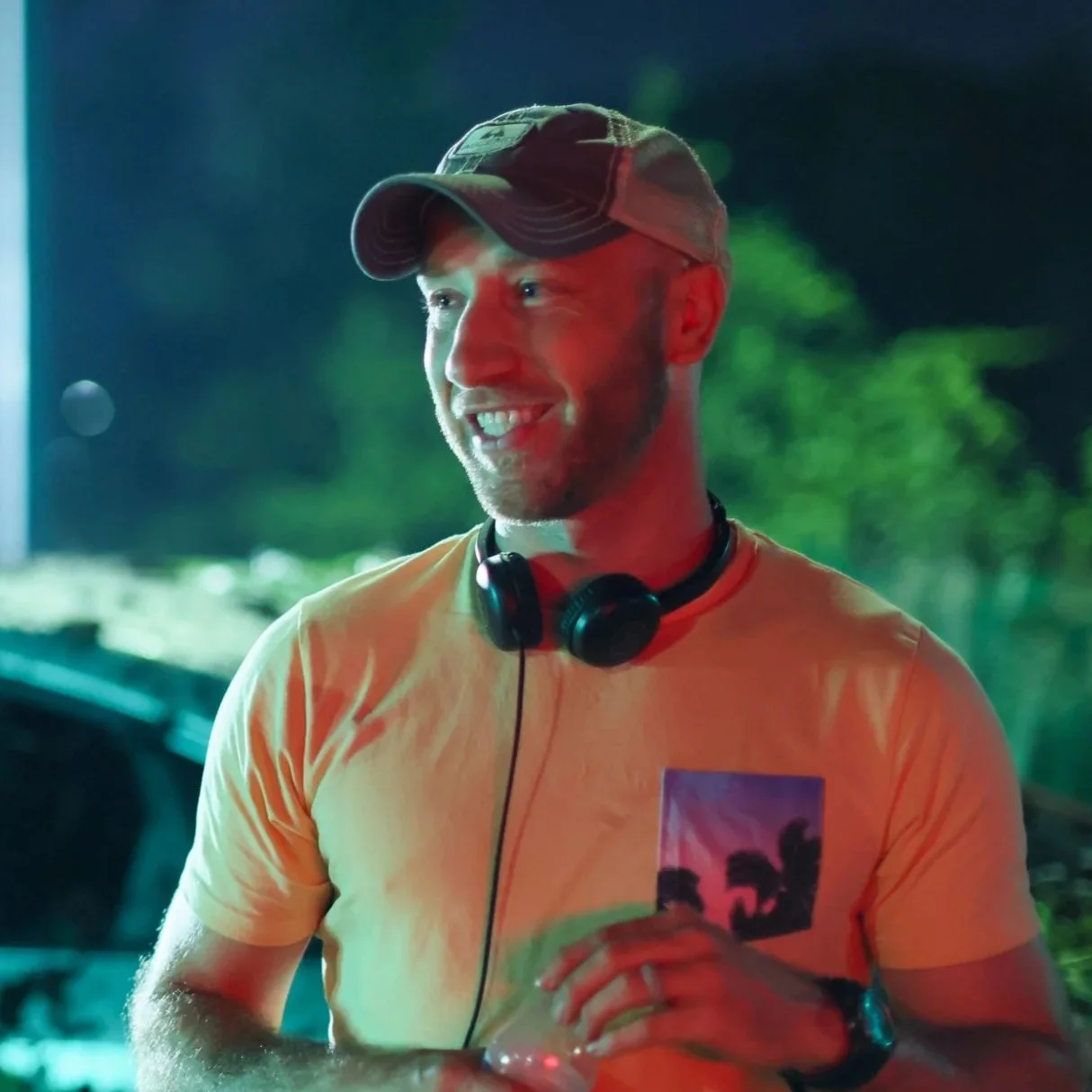Writer and director, Mitch Bechtold wearing a baseball cap and headphones around his neck, standing on the film set of his short film A Case For Coping, at night with green and red lighting effects.