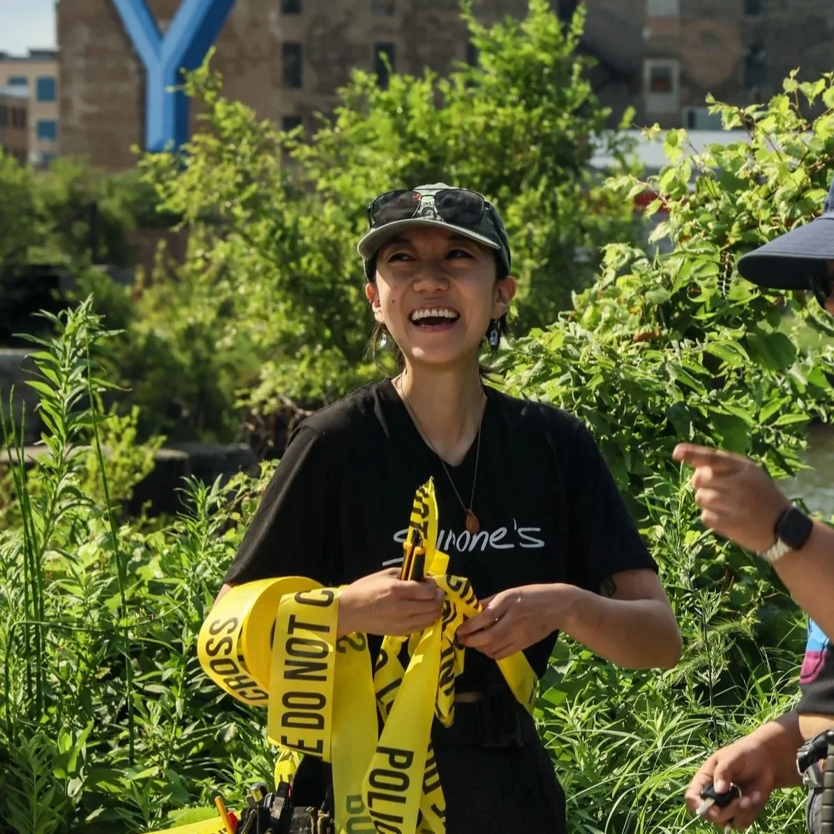 Production Designer, Linda Lee smiling and dressing the outdoors film set, wearing sunglasses, a grey cap, and black clothing, holding yellow caution tape with 'DO NOT CROSS' written on it, surrounded by green plants and trees.