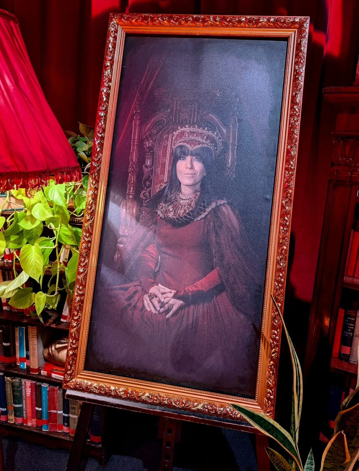 A framed portrait of a woman in historical clothing, sitting on a throne-like chair, with a red and gold ornate background. The portrait is displayed on an easel, to the left of a bookshelf with books and a green plant.