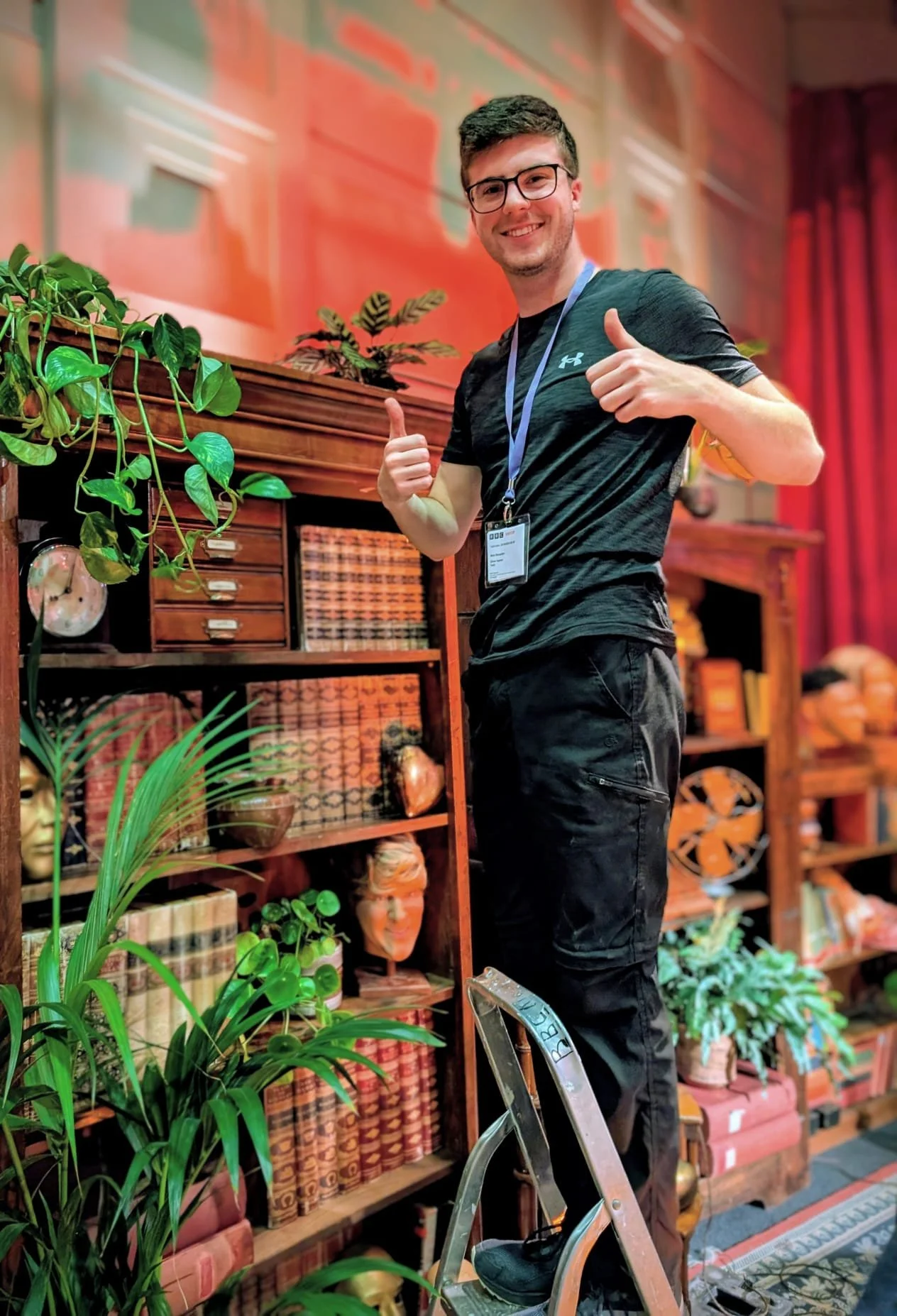 A young man with glasses and dark hair, wearing a black Under Armour t-shirt and black cargo pants, stands on a ladder giving a thumbs-up in a room decorated with plants, books, and decorative objects.