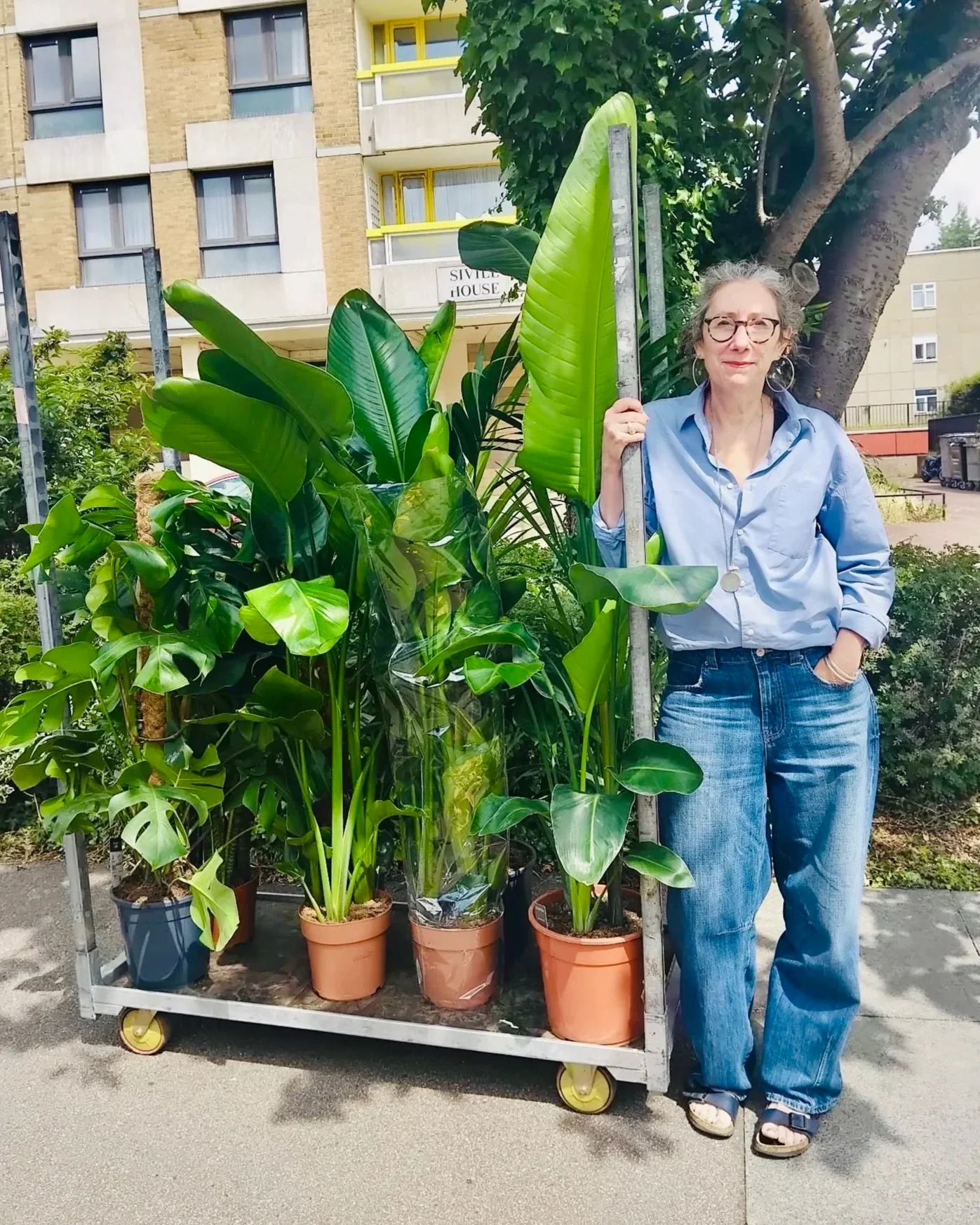 A woman standing next to a cart filled with large potted tropical plants and greenery on a sidewalk