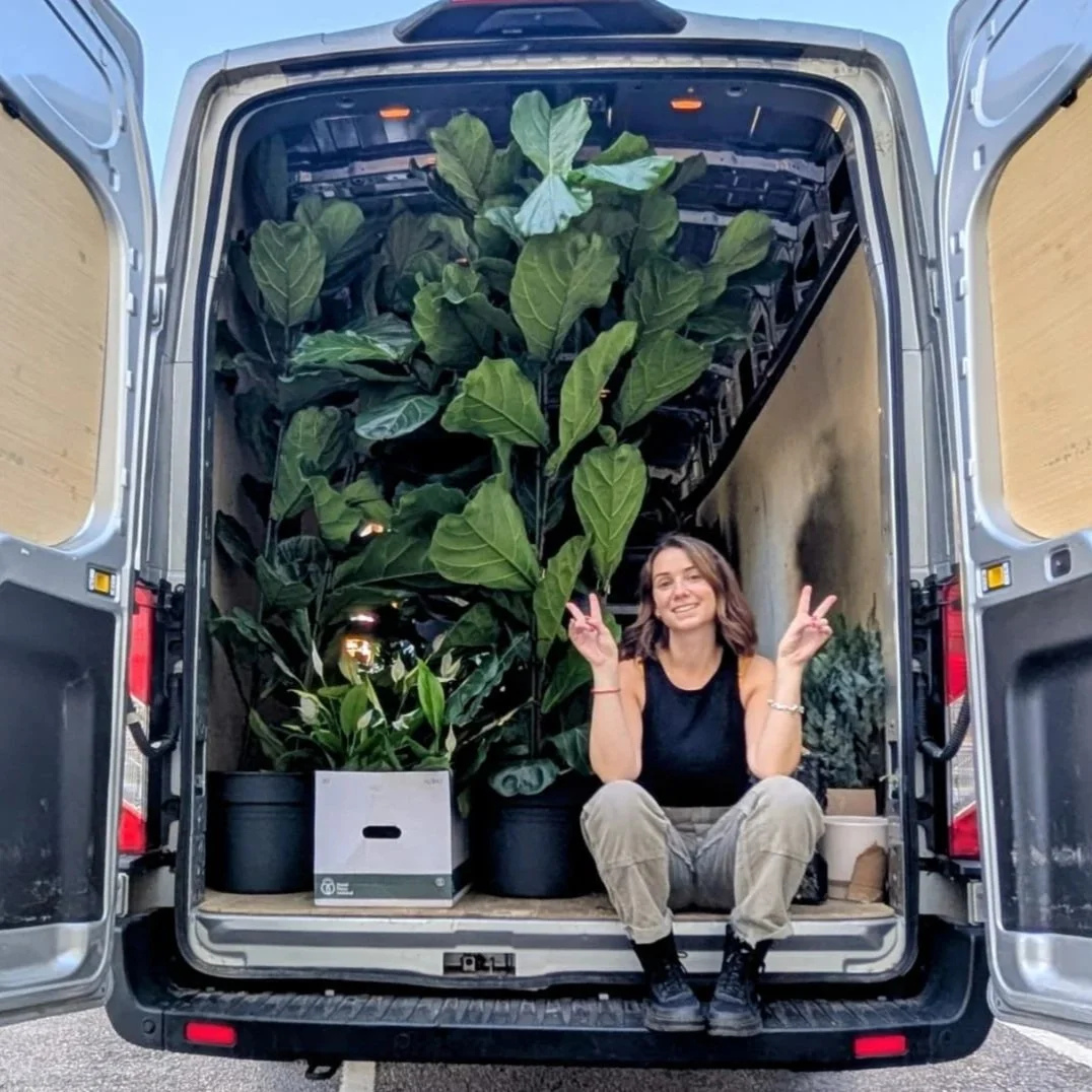 A young woman sitting inside the open back of a van filled with large potted green plants, making peace signs with both hands and smiling.