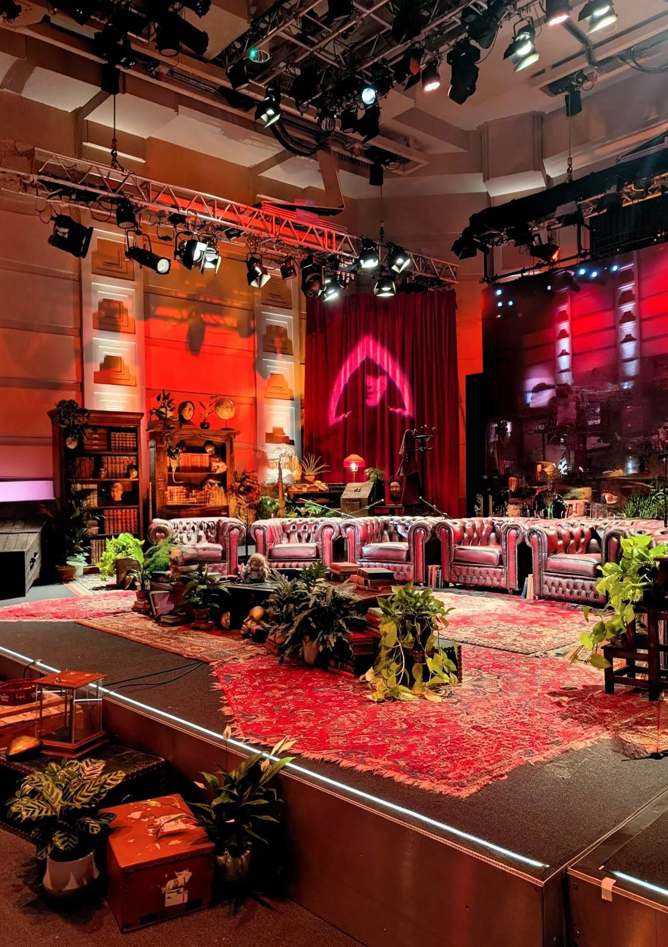 A stage set for a live TV show or performance, with vintage furniture including pink leather sofas, potted plants, bookshelves, and decorative items; red curtains and a neon pink triangle logo in the background; stage lighting and equipment overhead.