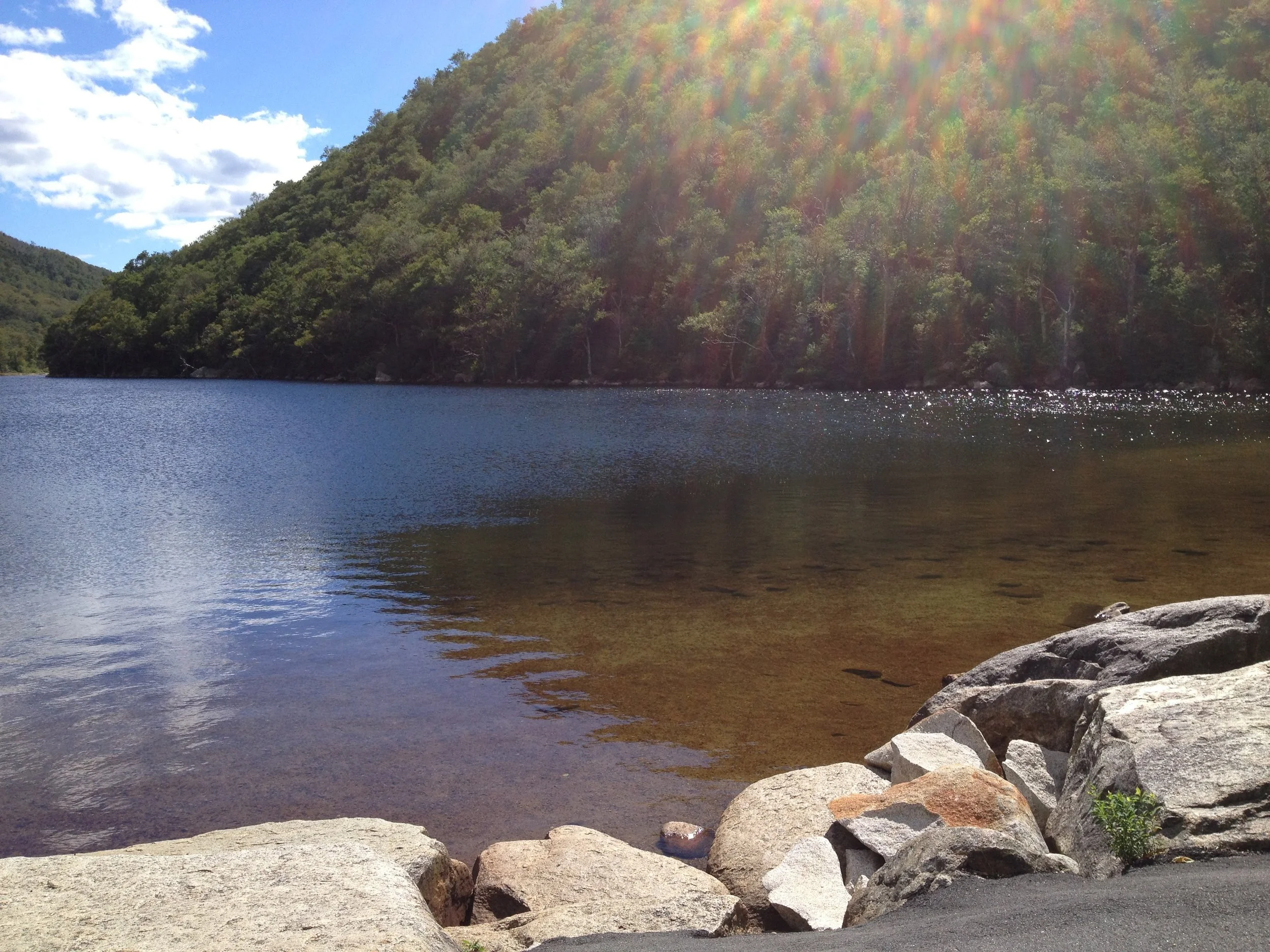 A lakeside scene with a rocky shore, calm water, wooded mountains in the background, a partly cloudy sky, and sunlight creating rainbow-like rays.