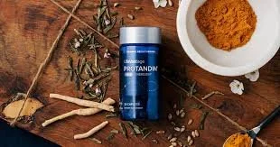Blue bottle of Protandim supplement on a wooden surface with herbal plants, small white flowers, a dish of turmeric powder, and mortar and pestle.