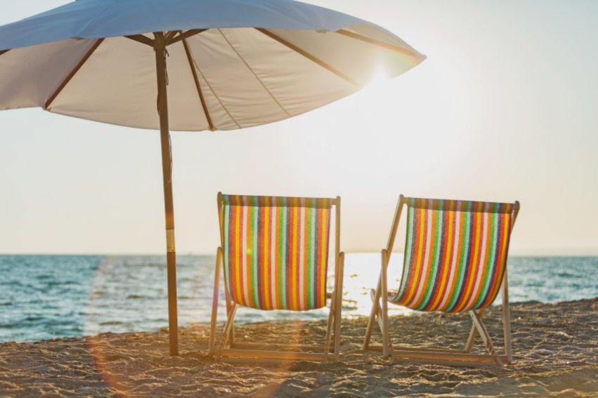 Two colorful striped beach chairs and a large beige umbrella on a sandy beach near the ocean at sunset.