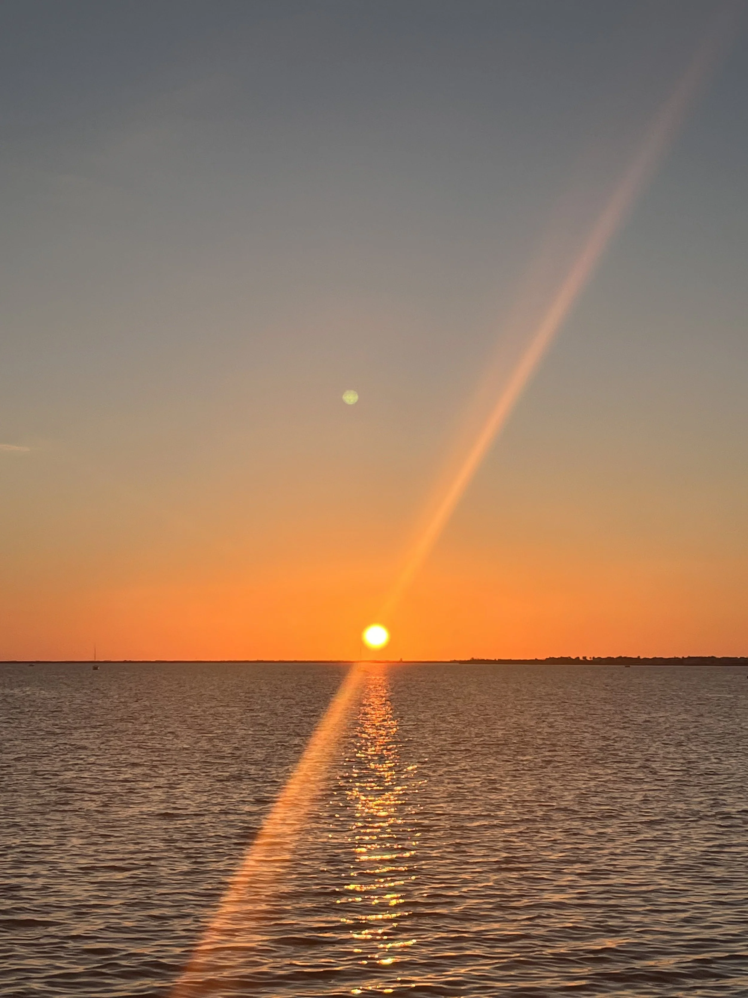 A sunset over a body of water with the sun near the horizon, creating a reflection on the water's surface, and a rainbow extending from the sky to the horizon.