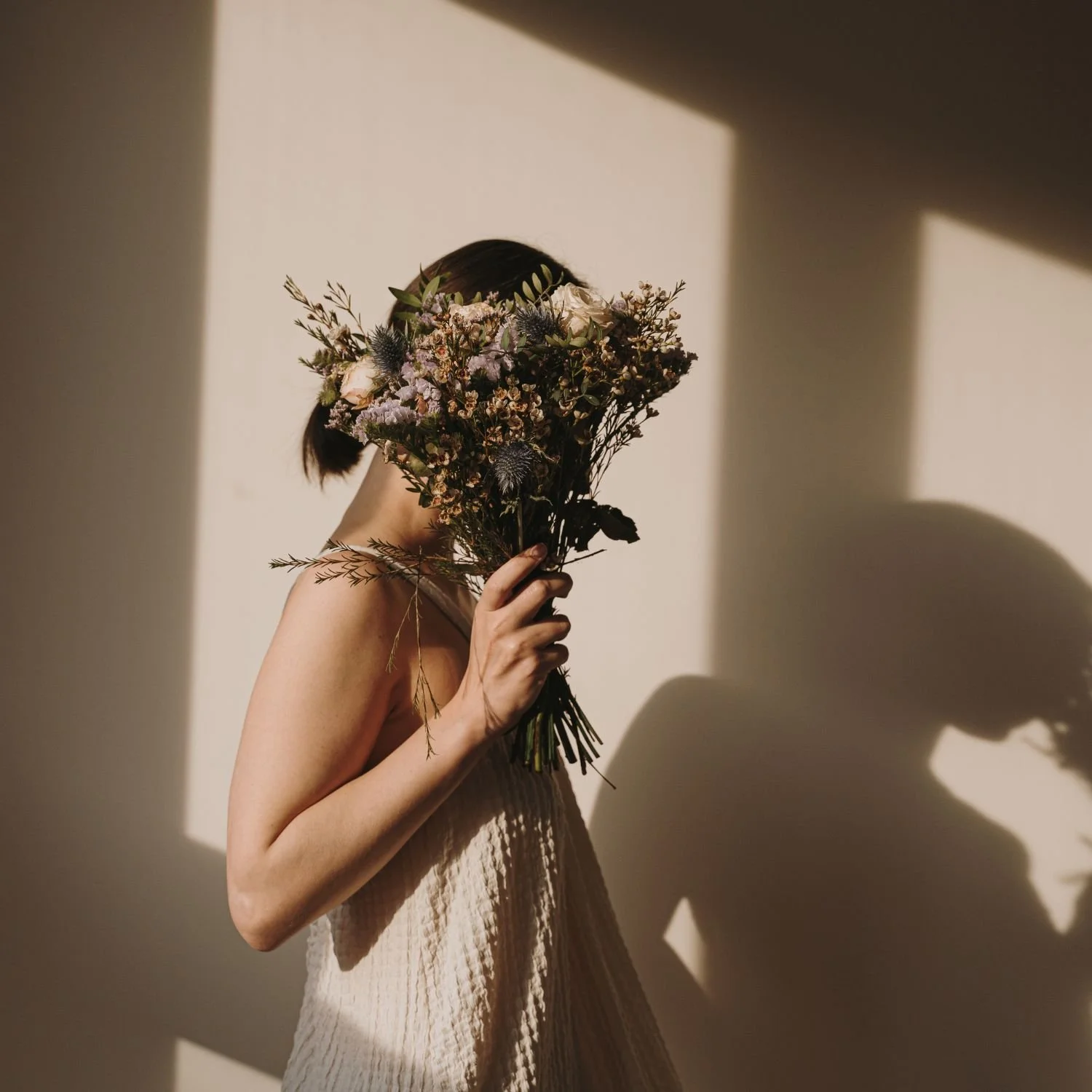 A woman in a light-colored dress holds a large bouquet of flowers in front of her face, casting a shadow on the wall behind her.