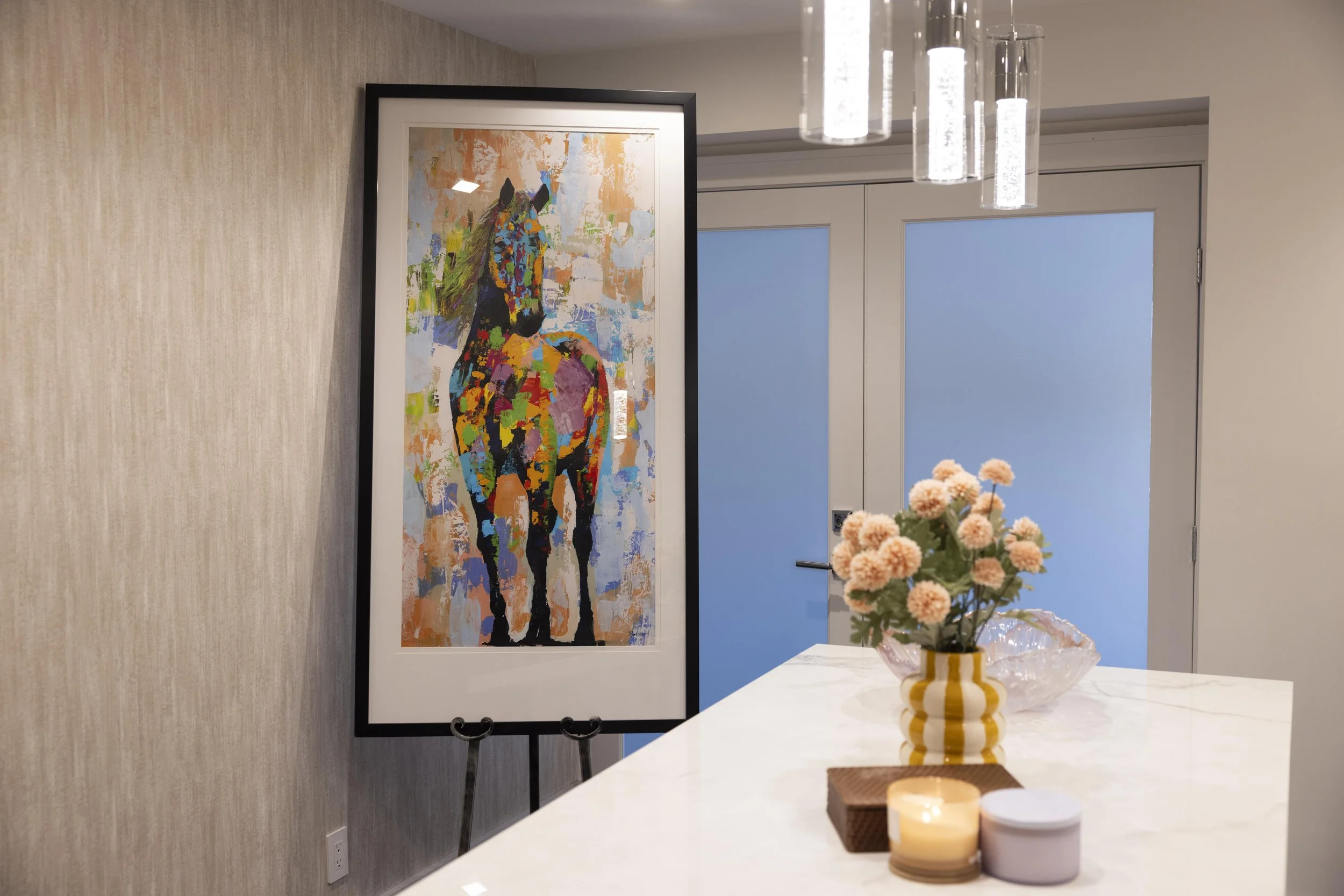 Interior of a modern room at Haus of Mental Health Counseling Long Island with a colorful horse painting on an easel, a vase with pinkish flowers, candles, and decorative items on a white table, with frosted glass sliding doors in the background.