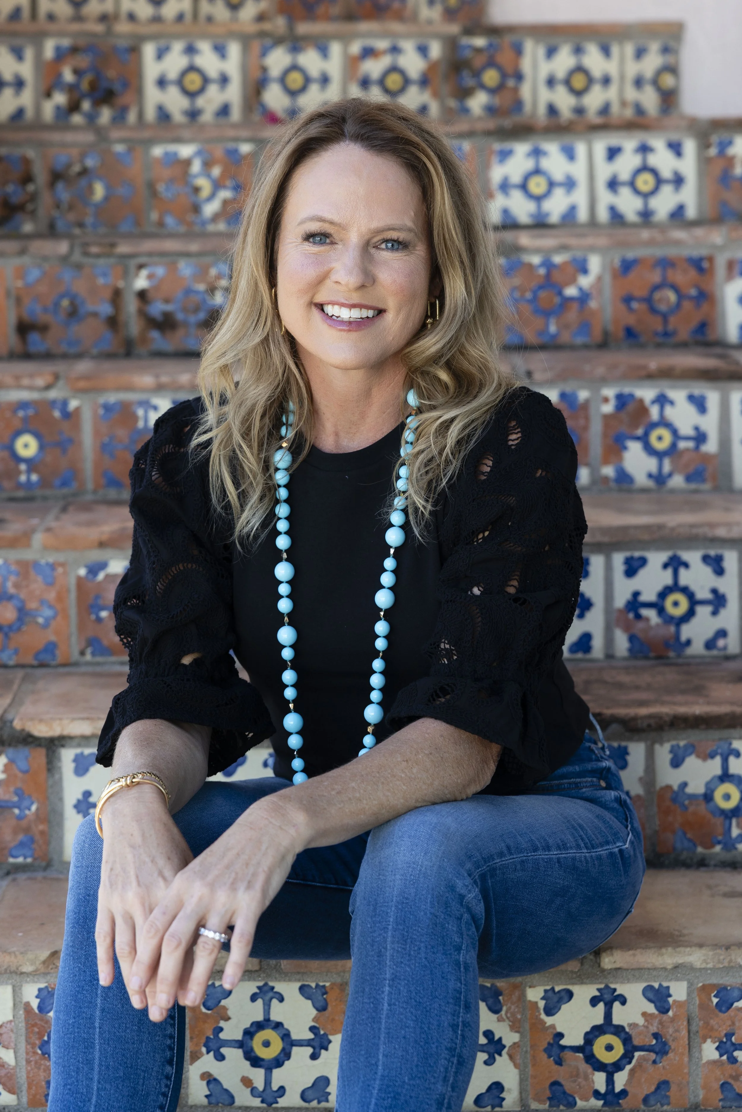 A woman with blonde hair, wearing a black top with lace sleeves, a turquoise beaded necklace, and blue jeans, sitting on tiled stairs with a colorful blue and white patterned background.