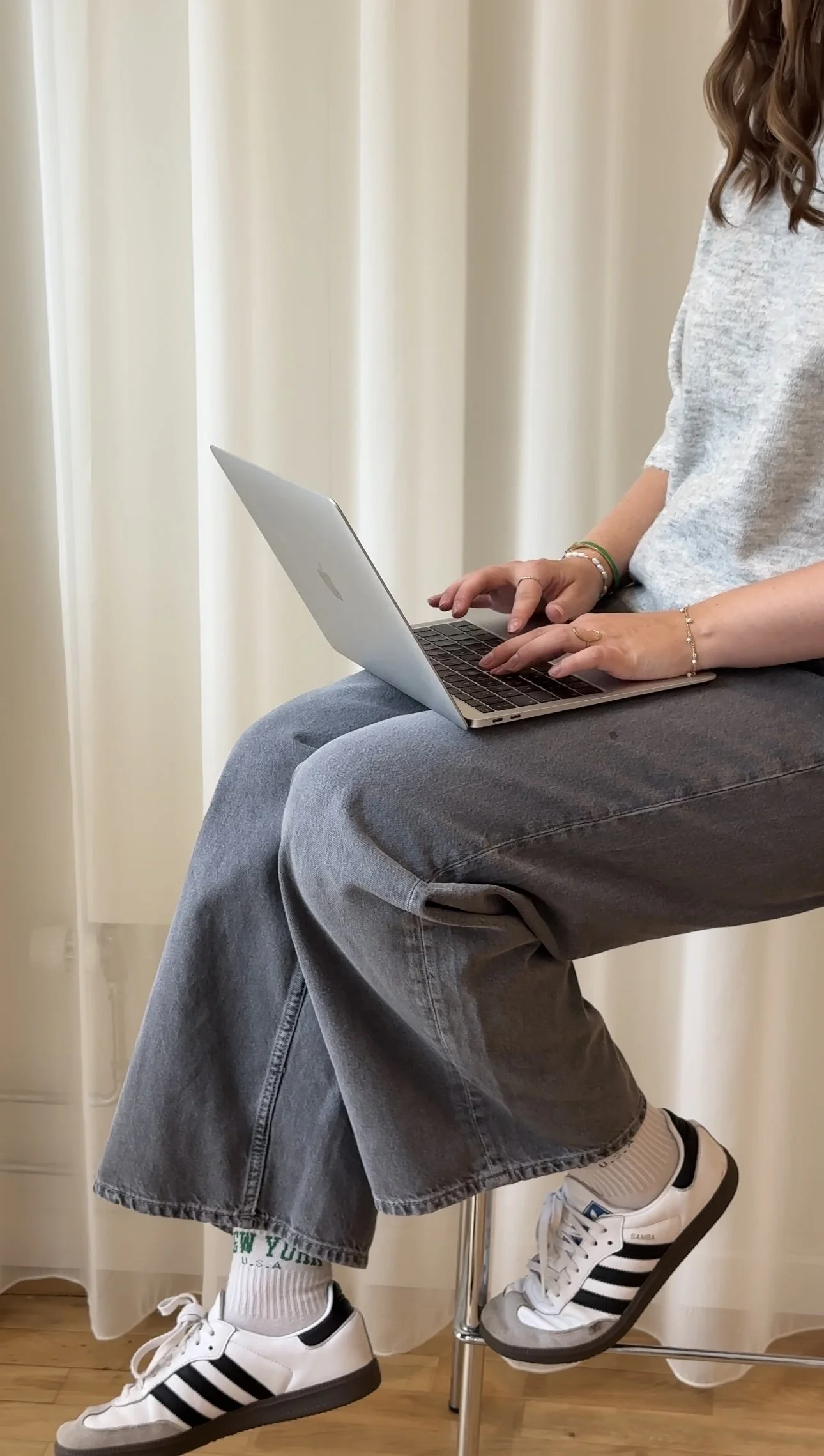 Person sitting on a high stool using a silver laptop. They are wearing casual gray jeans, a light gray shirt, white sneakers with black stripes, white socks, and have jewelry on their hands and wrists. The background has cream-colored curtains.