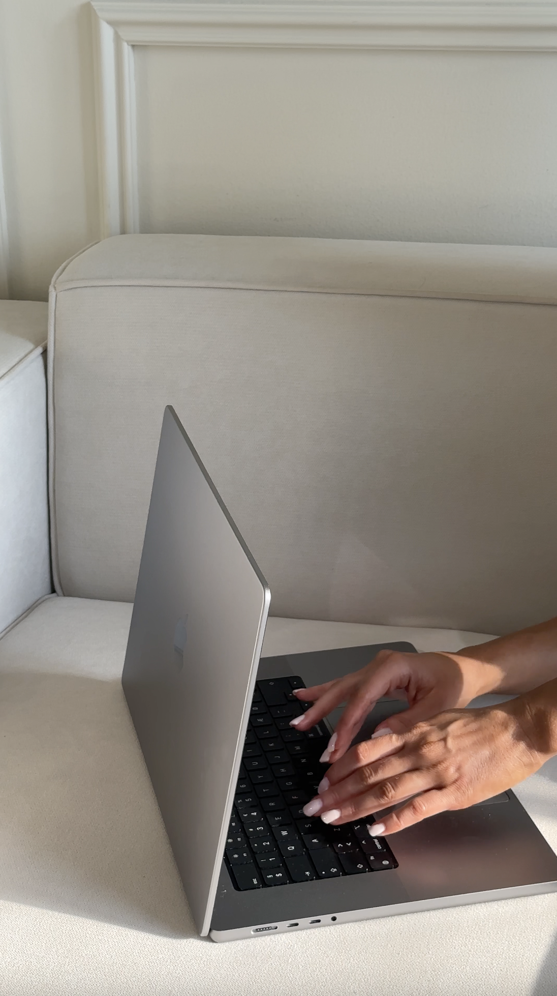 A person typing on a silver MacBook laptop with a black keyboard, sitting on a beige sofa, with part of their arm visible.
