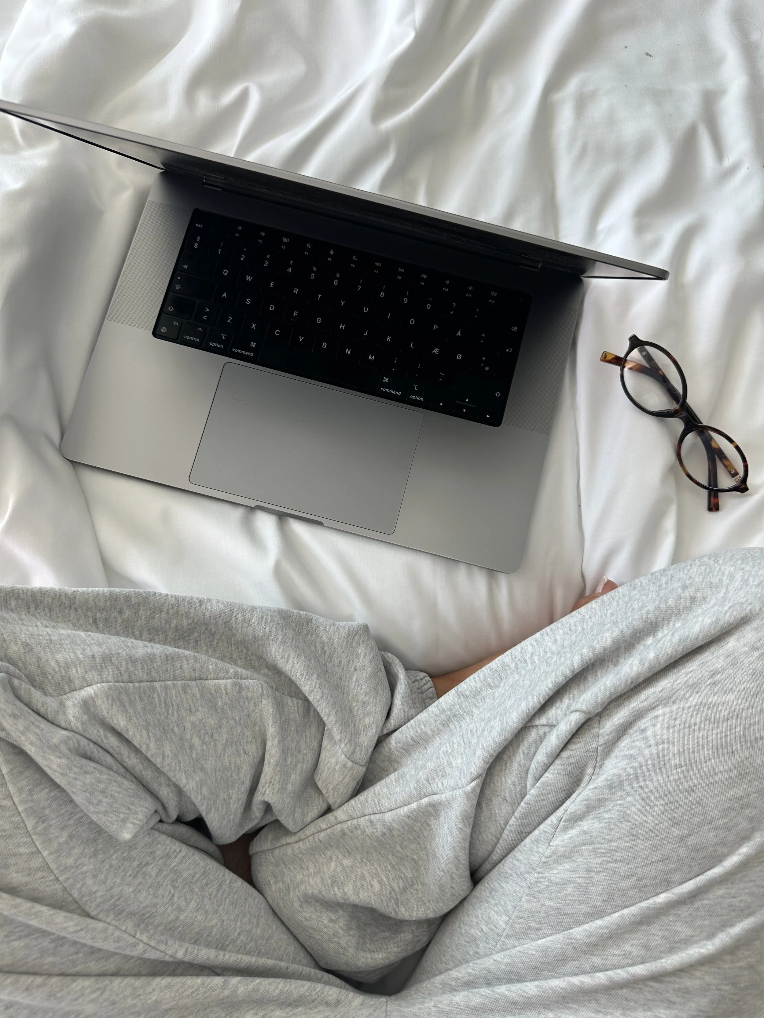 A person wearing light gray sweatpants sitting on white bed sheets, with a silver MacBook laptop and tortoise shell glasses nearby.
