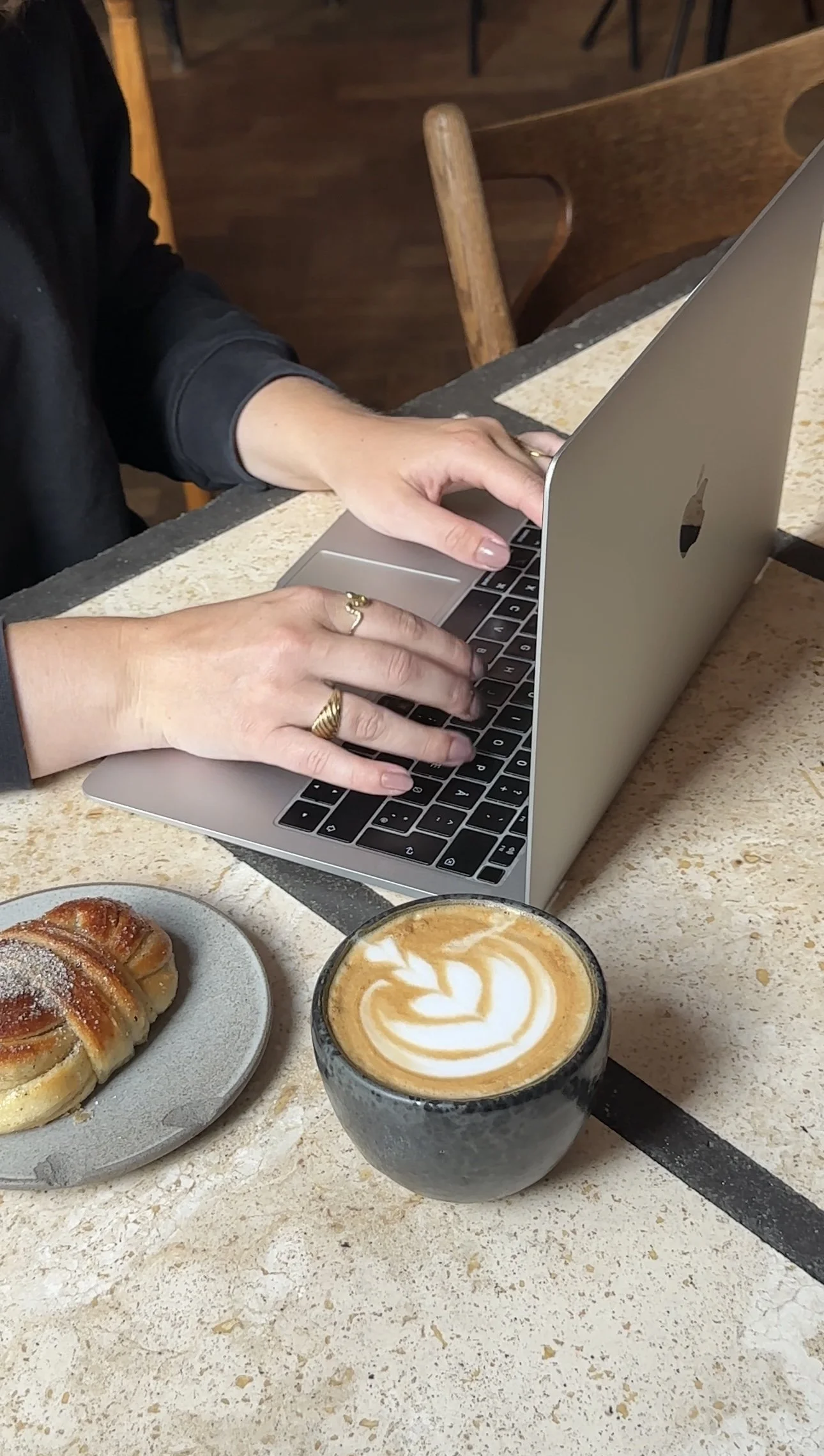 A person using a silver laptop at a restaurant table. There are pastries and a latte with latte art on the table.