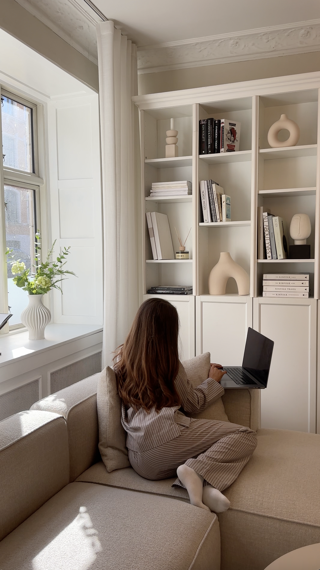 A woman with long brown hair wearing striped pajamas, sitting on a beige sectional sofa, using a laptop, in a bright living room with a large window with white curtains, a white bookshelf with decorative vases and books, and a white vase with green a