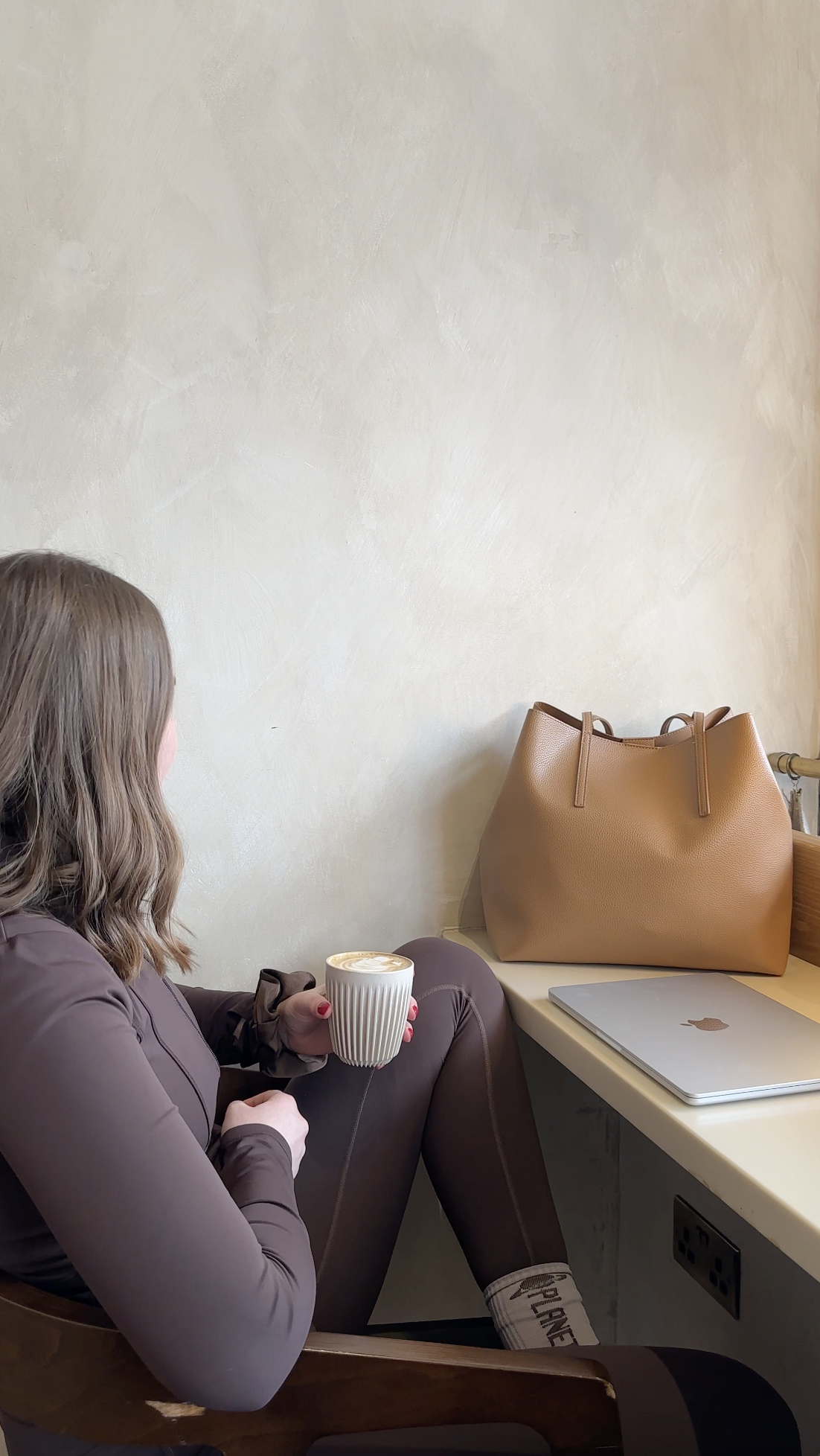 A woman with wavy hair sitting in a coffee shop, holding a cup of coffee, with a tan handbag and a laptop on the table beside her.