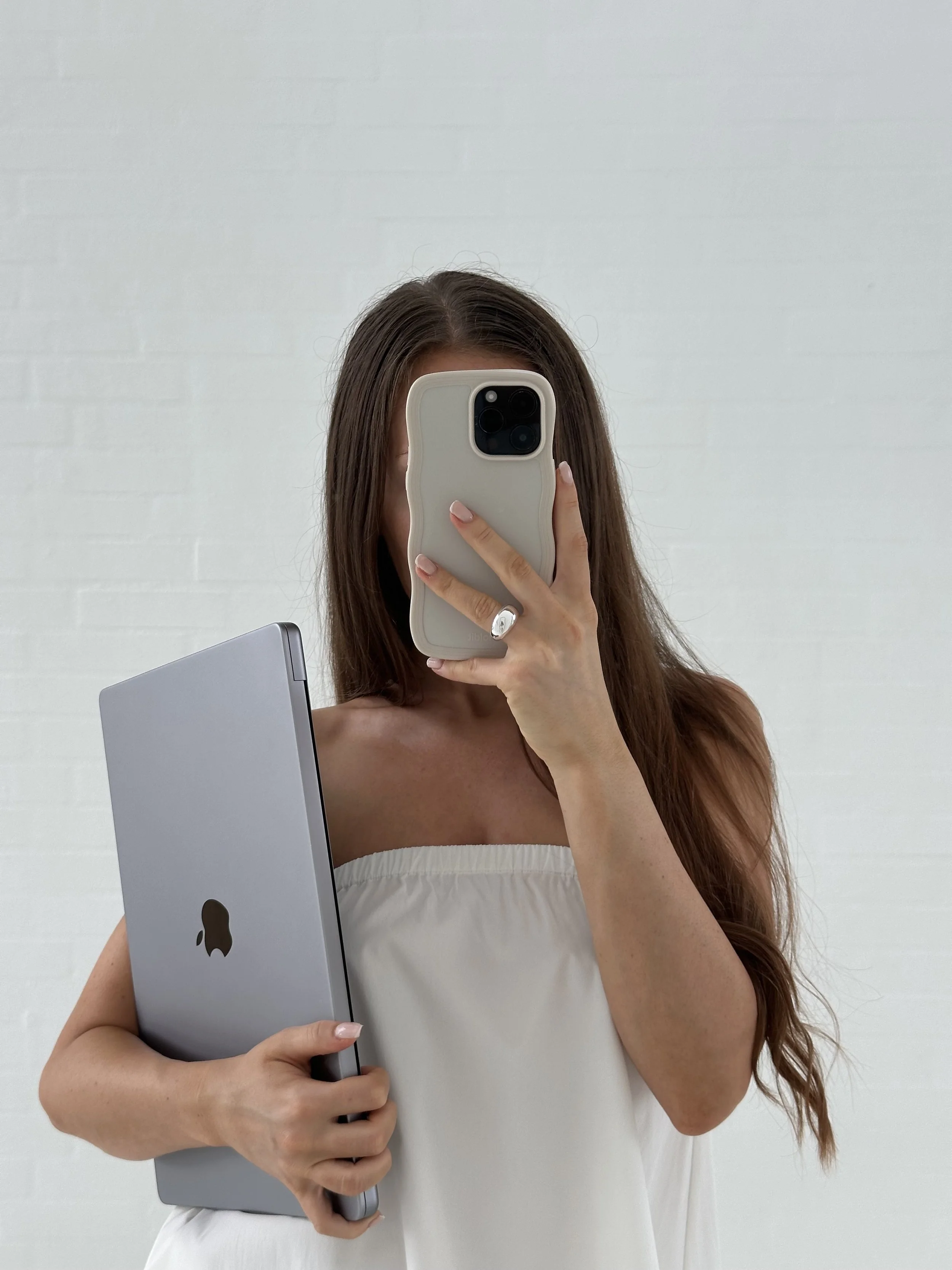 A woman with long brown hair is taking a mirror selfie with an iPhone, holding a silver laptop in her left hand, against a plain white wall background.