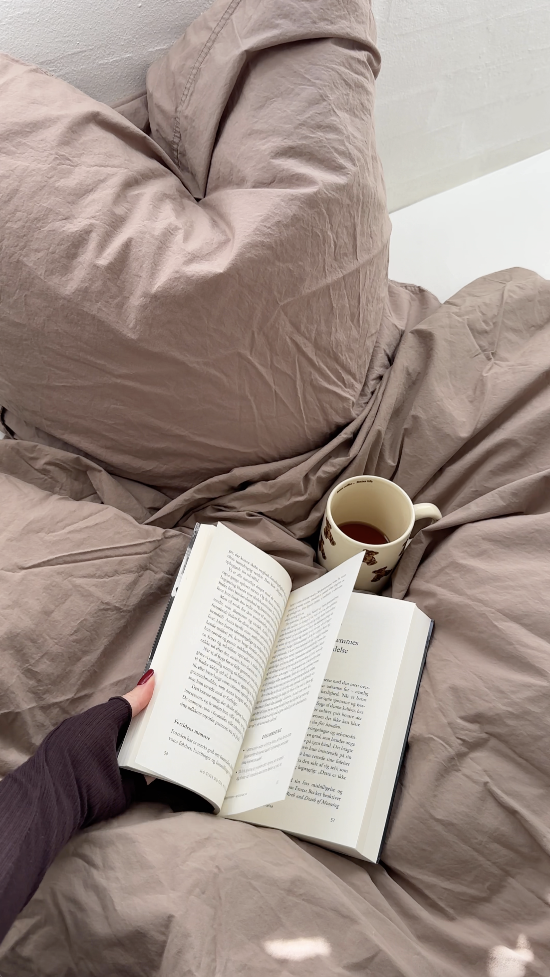 A person holding an open book while sitting on a bed with beige sheets. There is a mug with a floral pattern and a dark beverage on the bed. The person's hand has red nail polish and they are wearing a dark long-sleeve shirt.